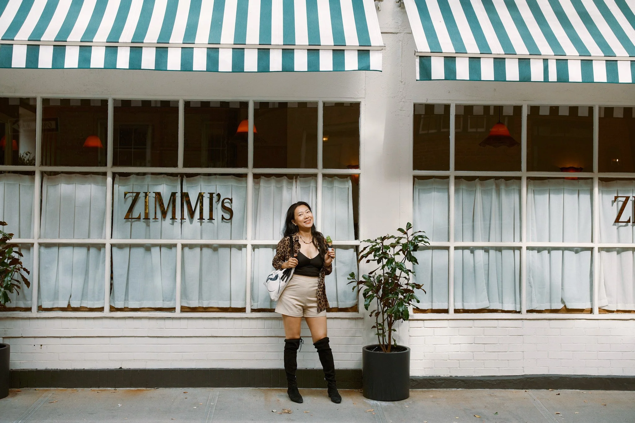 A woman posing outdoors in front of a large windowed building with striped awnings, a potted plant, and the partial signage 'ZIMMI'S'.