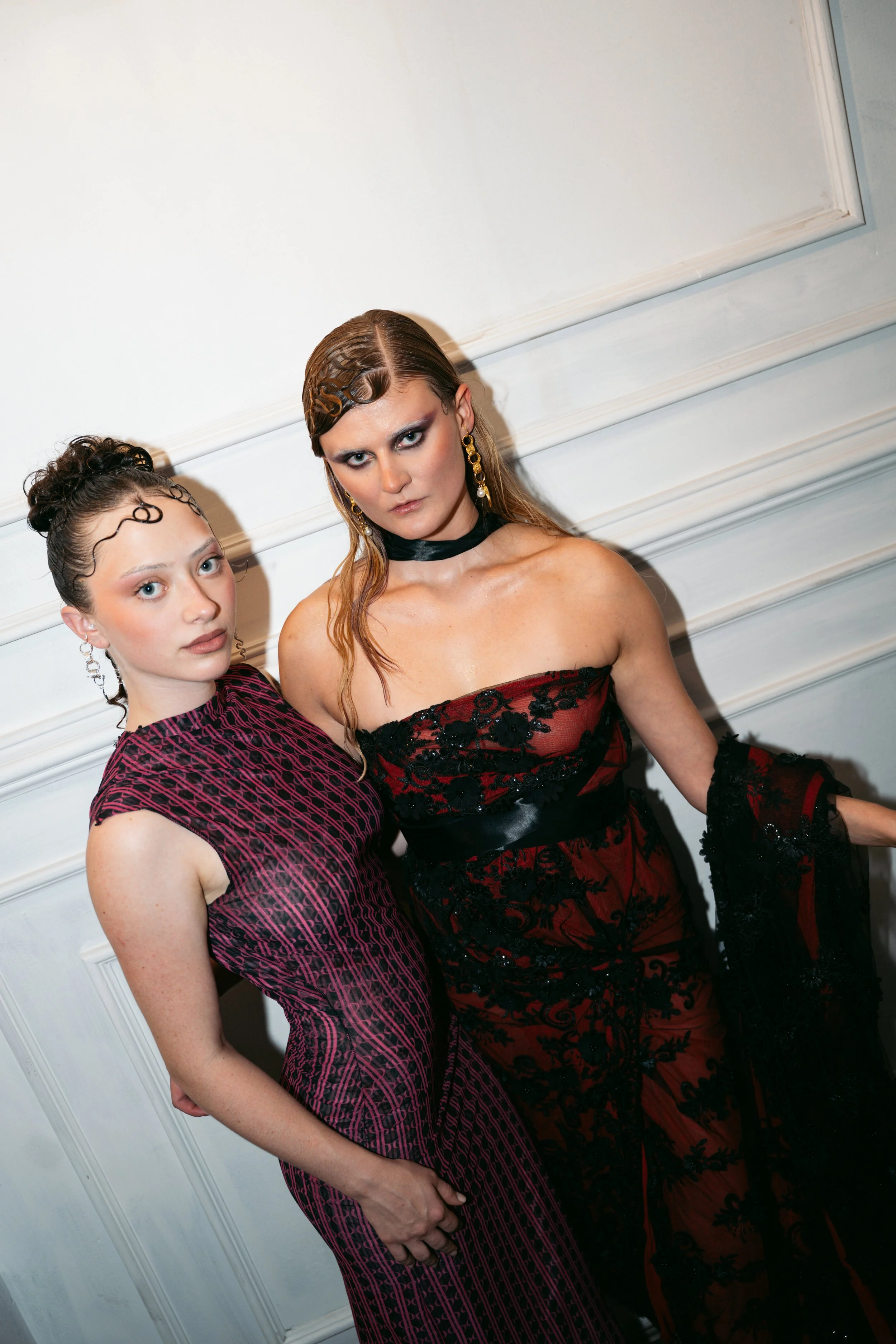 Two women dressed in elegant evening wear with intricate designs, standing against a white panel wall. One woman has curly hair styled up, and the other has long, wavy hair with makeup and earrings.