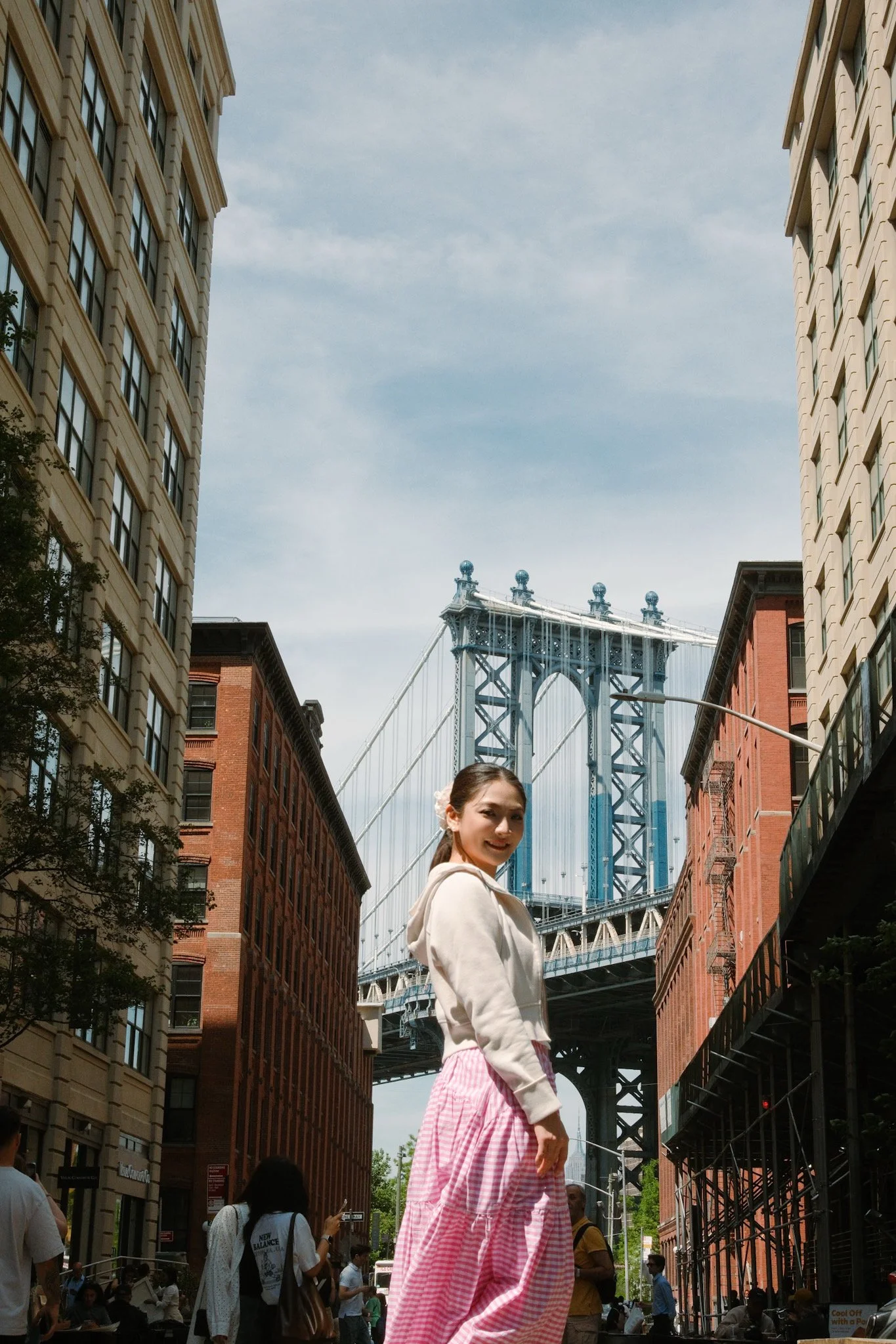 A young woman in a white hoodie and pink gingham skirt standing on a city street near the Manhattan Bridge in New York City, with buildings on either side and people walking in the background, under a partly cloudy sky.