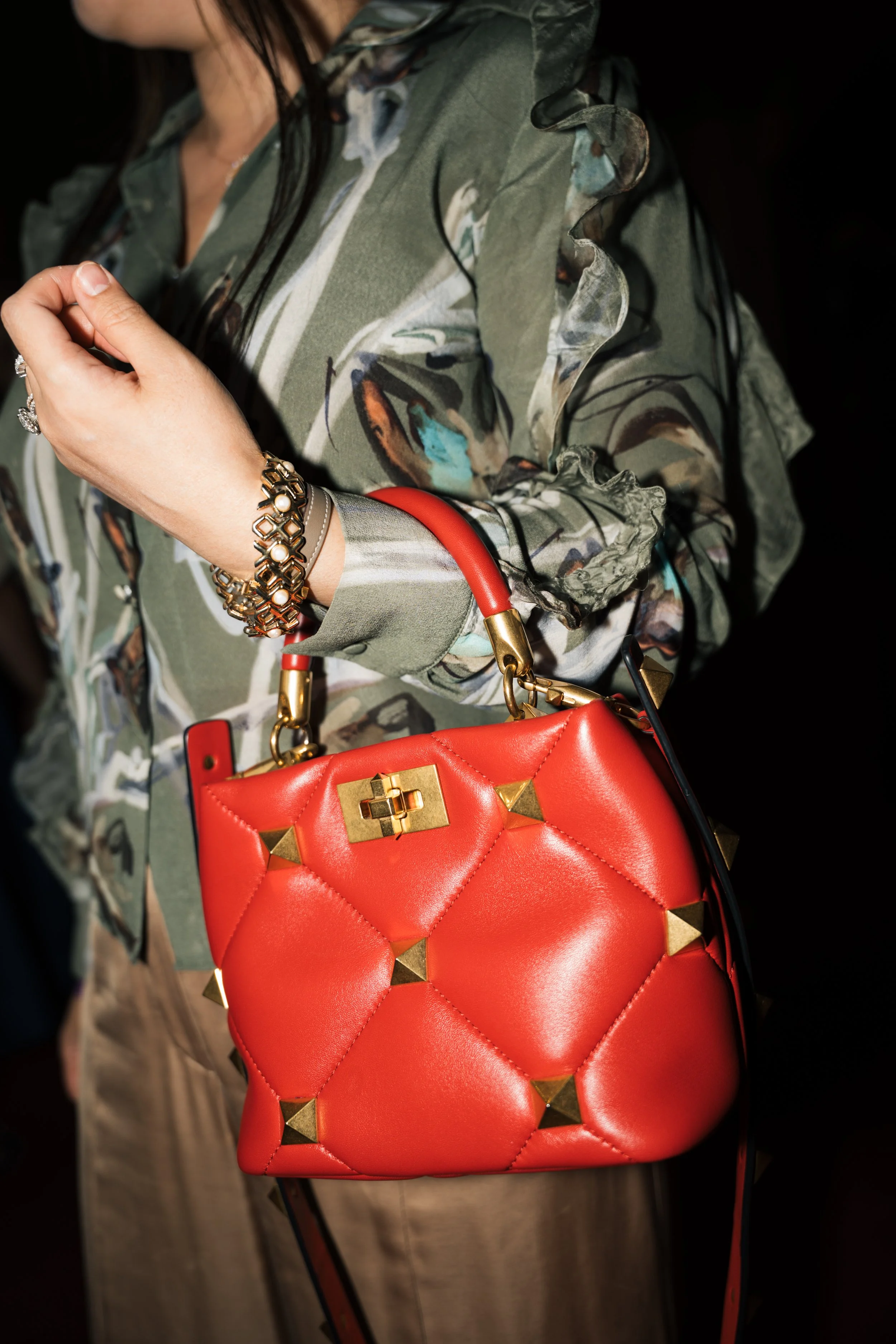 A woman wearing a green floral blouse with ruffled sleeves, holding a red quilted handbag with gold studs, gold hardware, and a red strap. She is also wearing a bracelet and a ring.