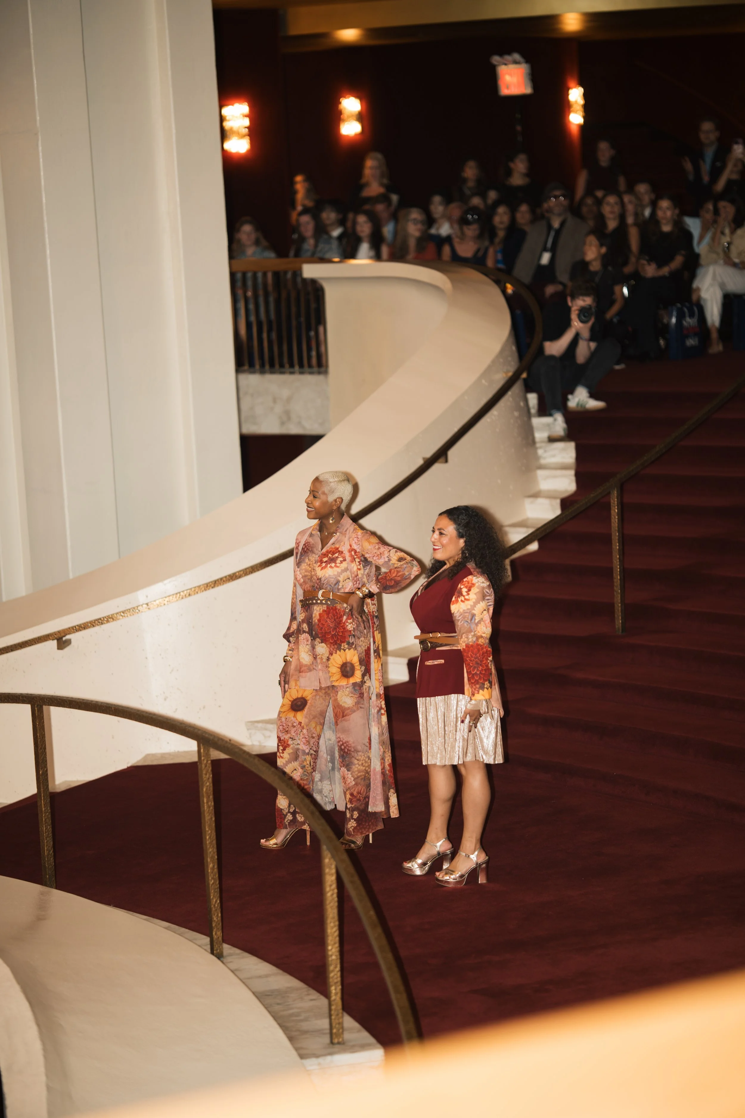 Two women standing on a staircase in an elegant venue, with an audience seated in the background. The women are smiling and dressed in colorful, patterned outfits with high heels.
