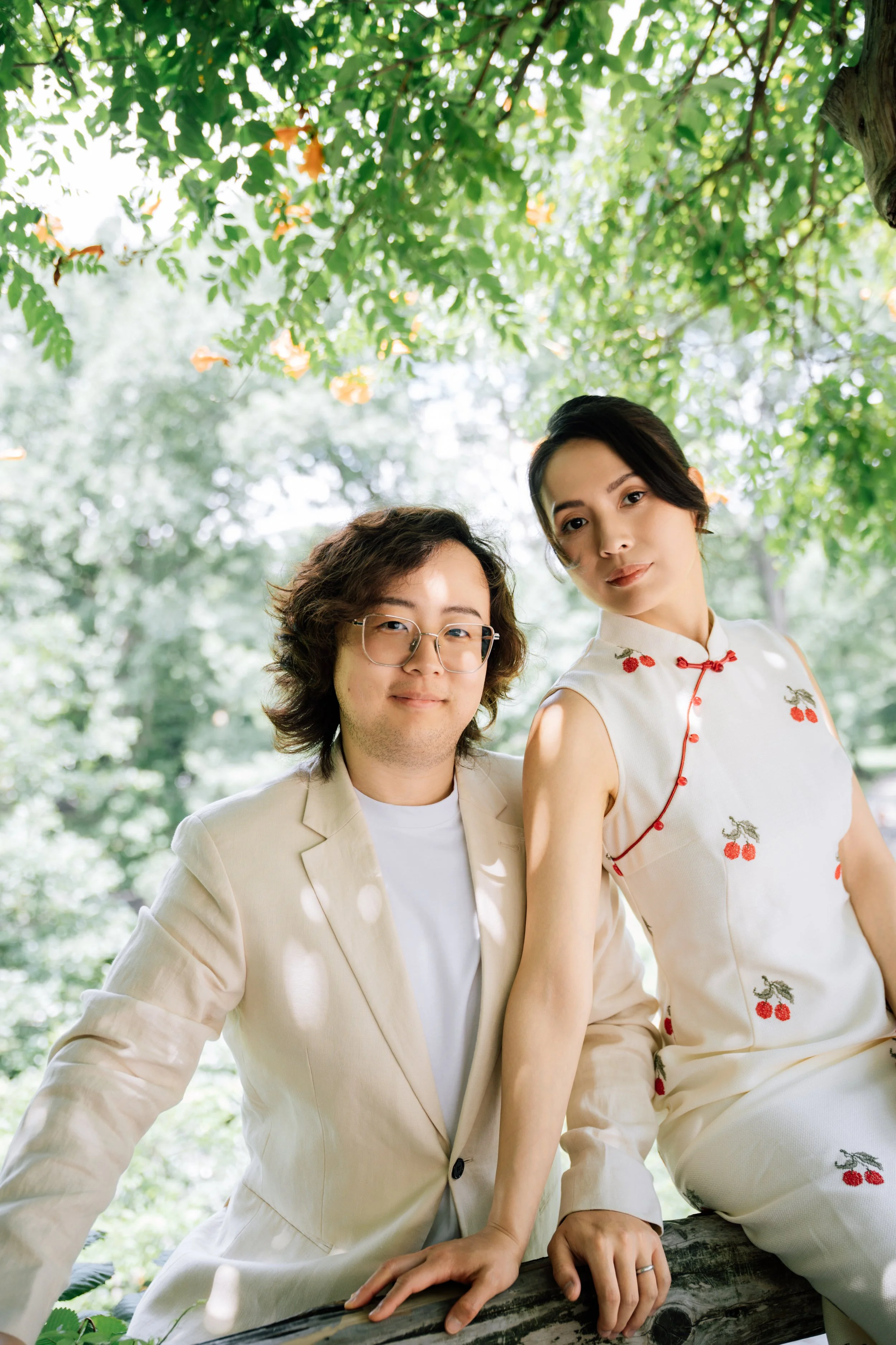 Two people, a man with curly hair and glasses, and a woman with short dark hair, pose outdoors under a leafy tree. The man wears a light beige suit jacket, and the woman wears a cream-colored dress with cherry and dragon embroidery and red trim.
