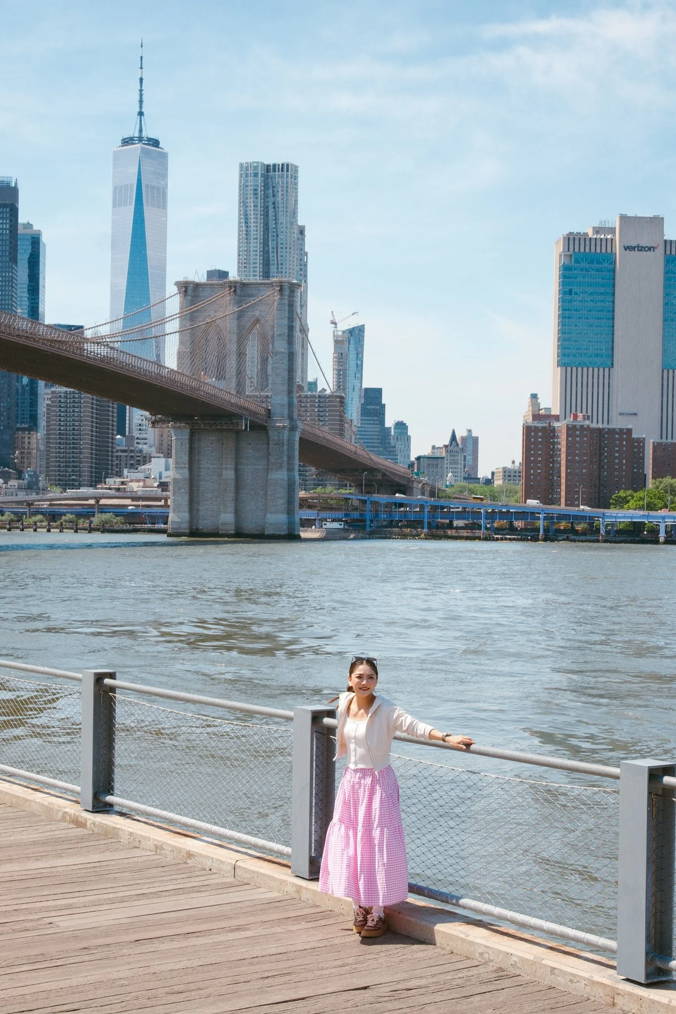 A young woman in a pink and white checkered skirt and white top standing near a waterfront railing with the New York City skyline, including the Brooklyn Bridge and tall skyscrapers, in the background.