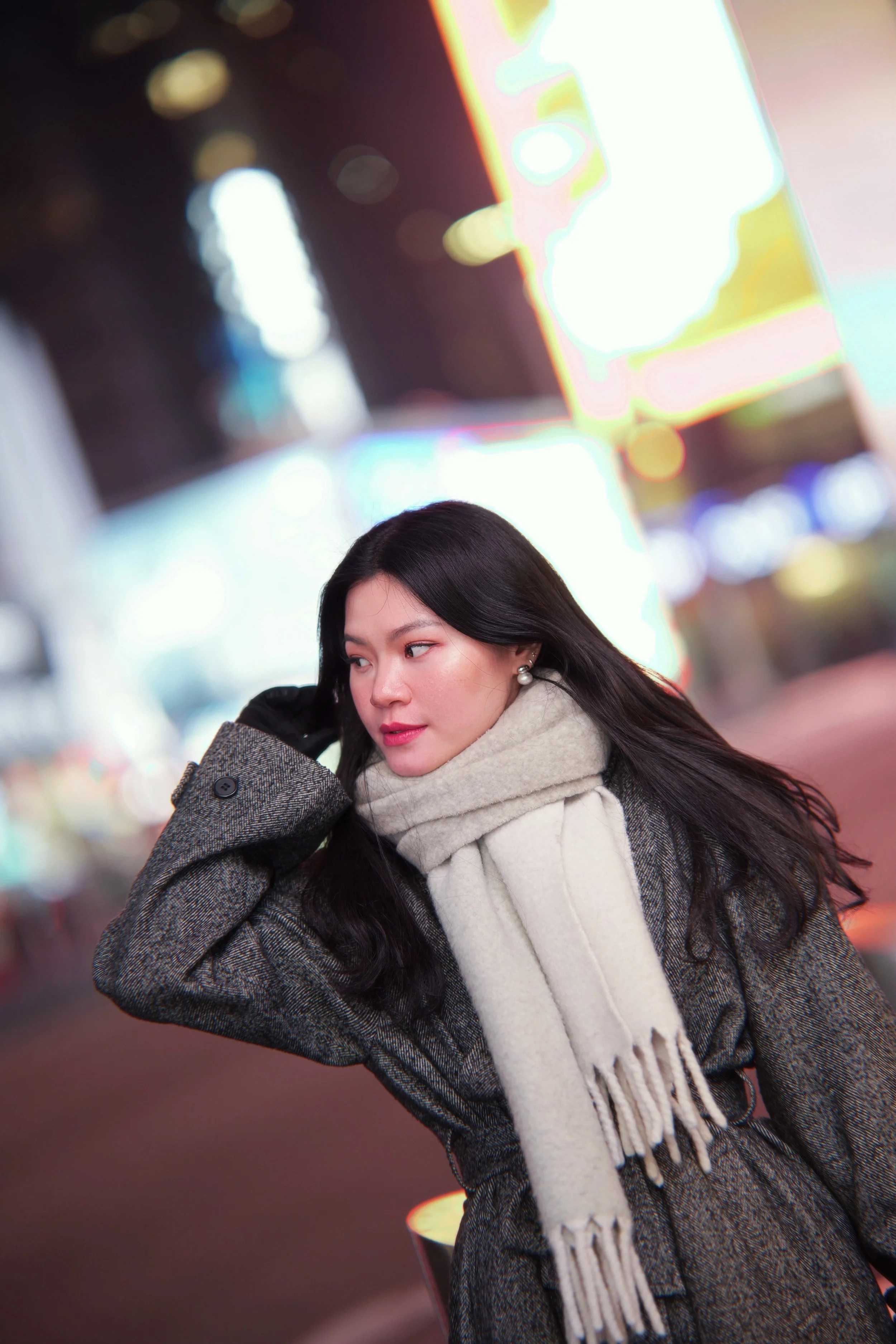 A young woman with long black hair and pearl earrings standing on a city street at night, wearing a gray coat and beige scarf with fringe, illuminated by colorful city lights.