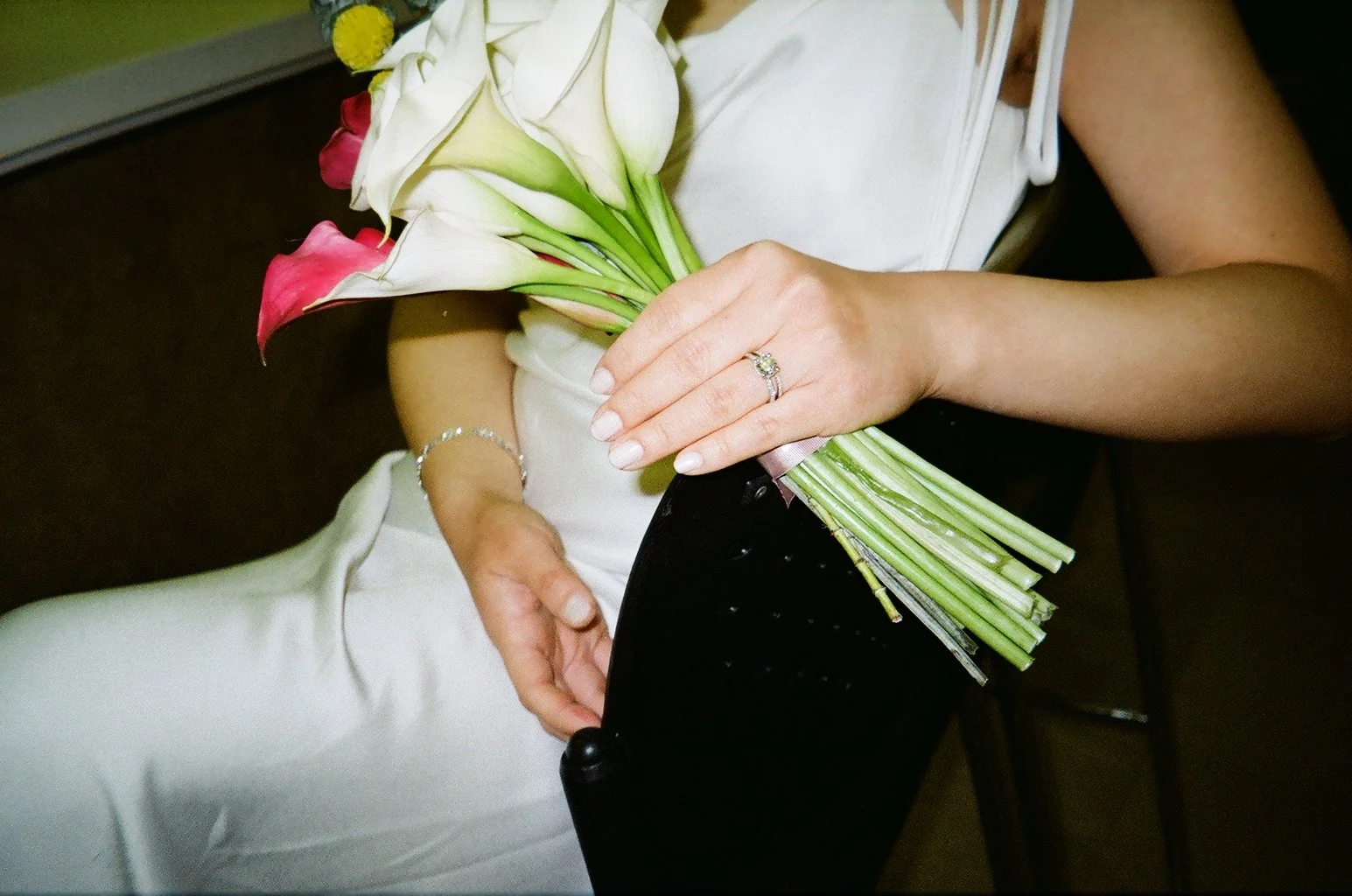 A woman holding a bouquet of white and pink calla lilies, wearing a white dress, with a ring and bracelet on her hands, seated on a dark chair.