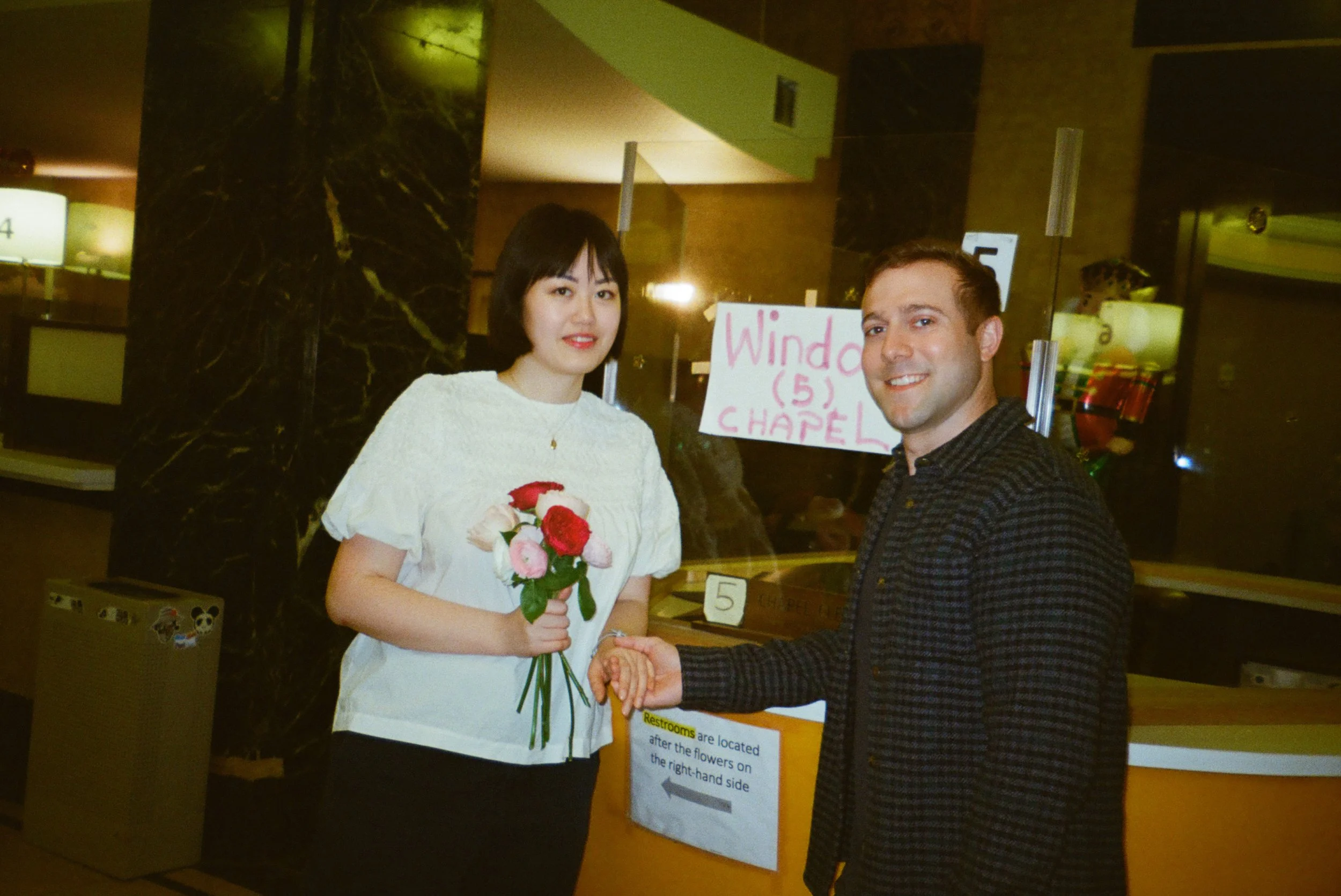 A woman holding a bouquet of flowers standing next to a man, shaking hands, in a lobby or reception area. There are signs and decorations in the background, indicating a public or event setting.