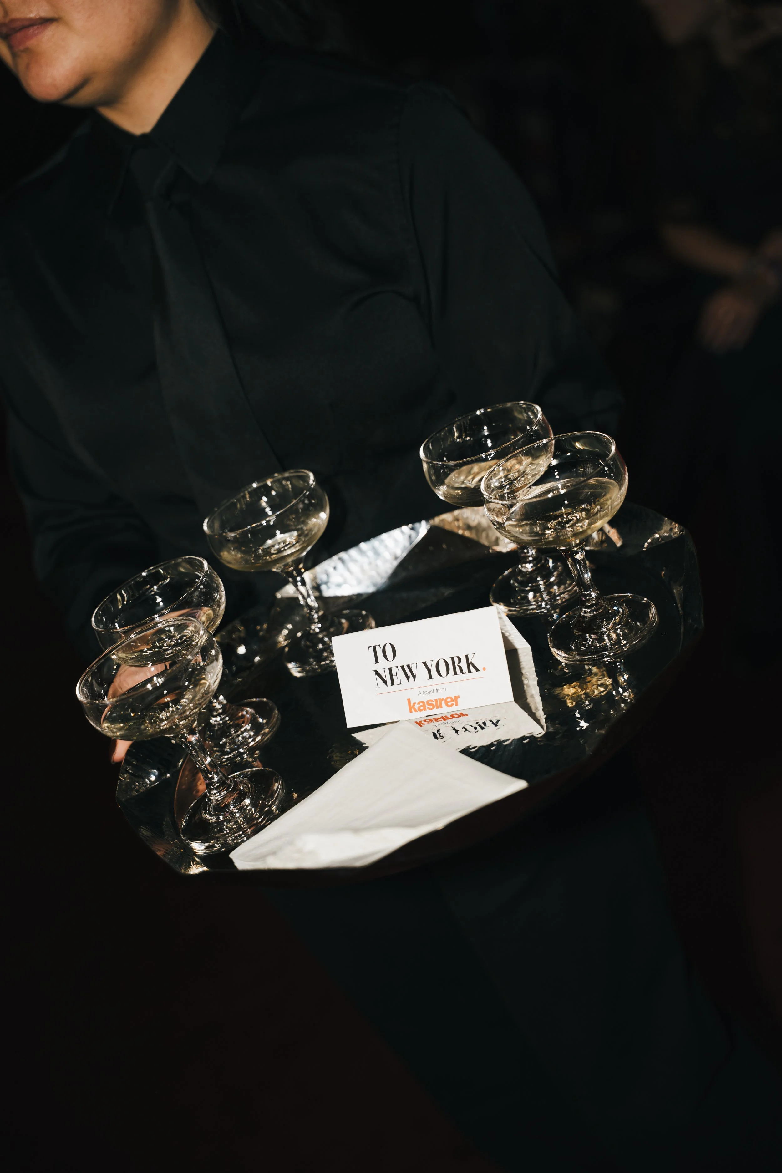 A person dressed in black attire is holding a tray with eleven champagne glasses filled with a sparkling beverage. There is a paper sign on the tray that says "TO NEW YORK" with the logo of Kasirer.