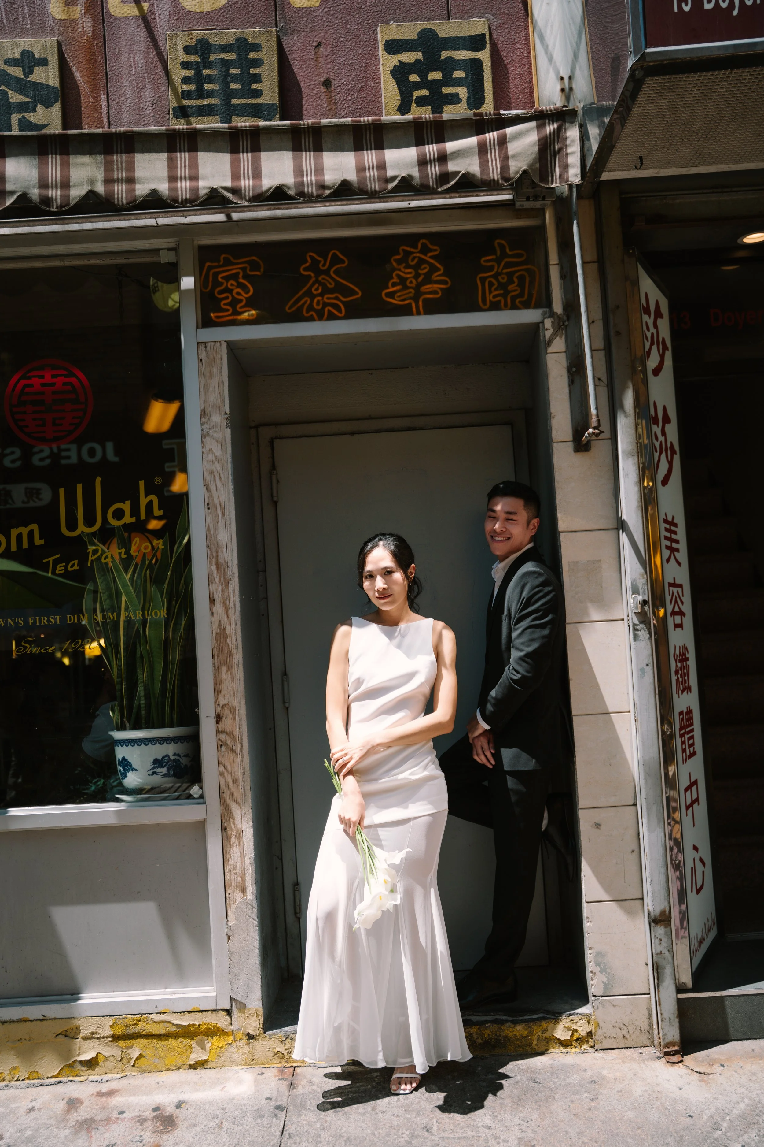 A woman in a white dress holding flowers and a man in a black suit jointly pose for a photo outside a restaurant with Asian signs. They are smiling and standing under a striped awning.