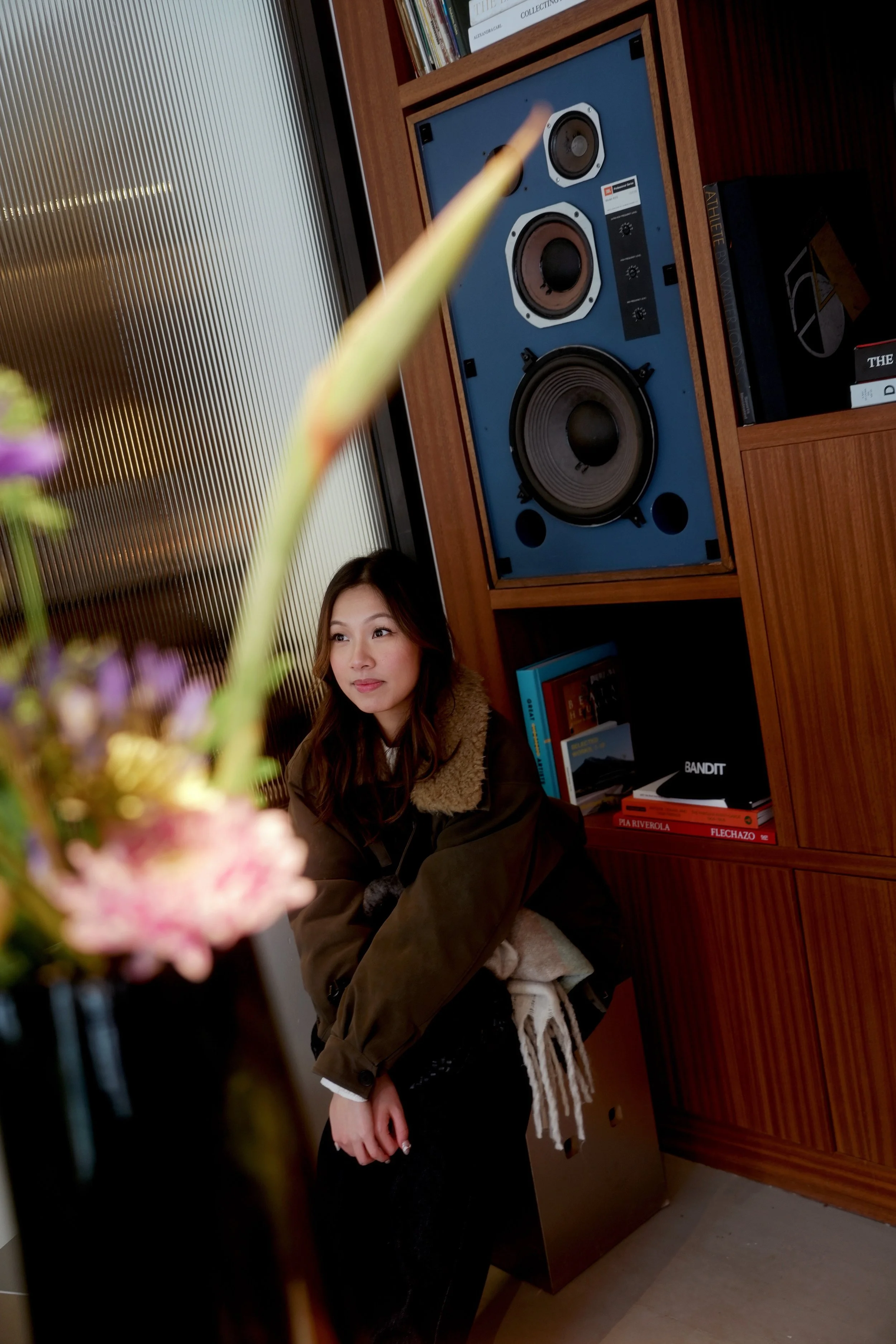 A young woman with long brown hair, wearing a brown jacket with a furry collar, sits in a room with wooden bookshelves, a colorful book collection, and a large blue and black speaker in the background. There are blurred flowers in the foreground.