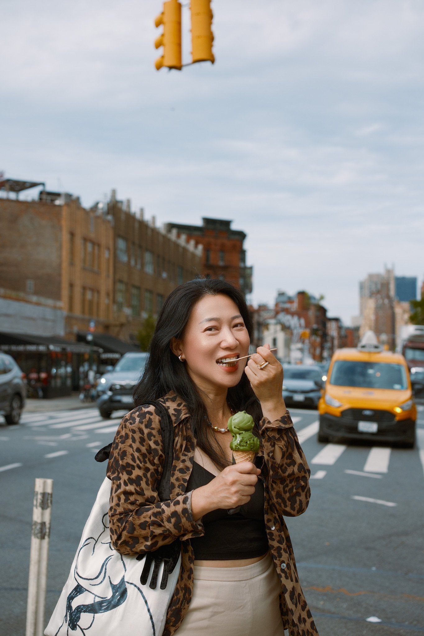 A woman with dark hair, wearing a leopard print top and beige skirt, is outdoors in an urban area, holding an ice cream cone with mint green ice cream, and smiling while looking at the camera. Behind her, there are cars, buildings, and a traffic ligh