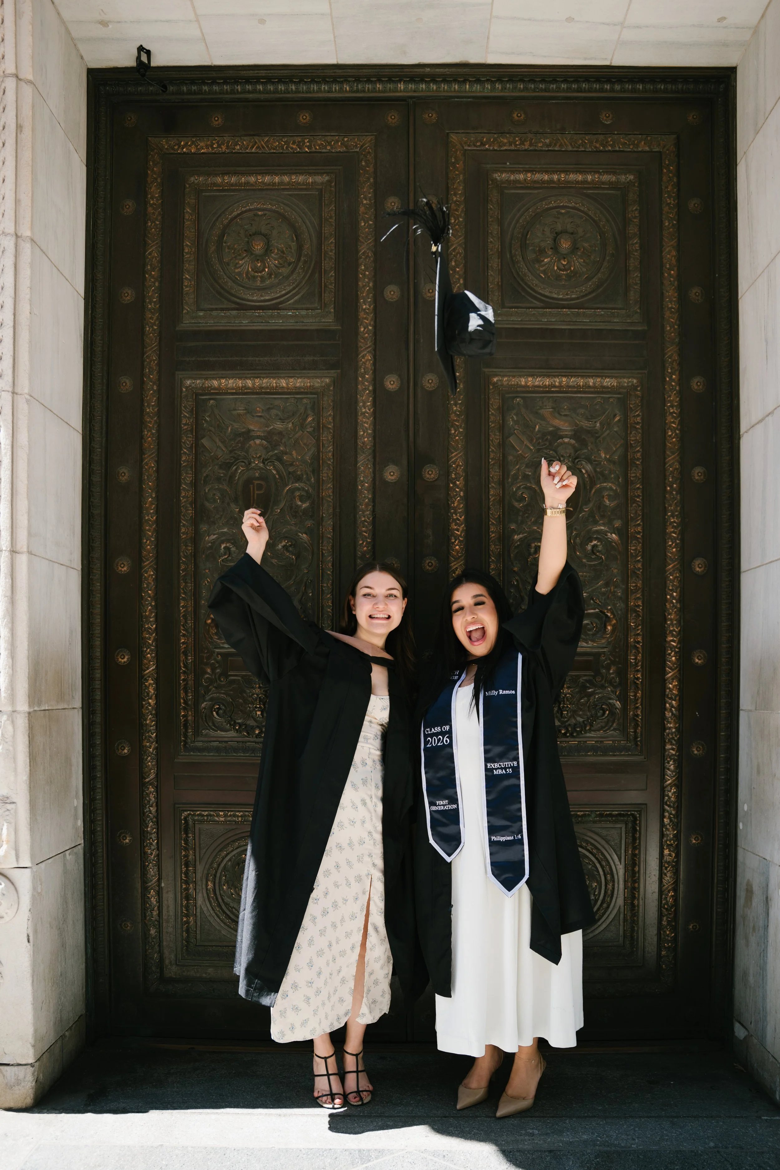 Two women in graduation gowns celebrating in front of a large ornate door. One woman is wearing a white dress with a floral pattern and black high heels, and the other is wearing a white dress with beige heels. Both are smiling, with one raising her 