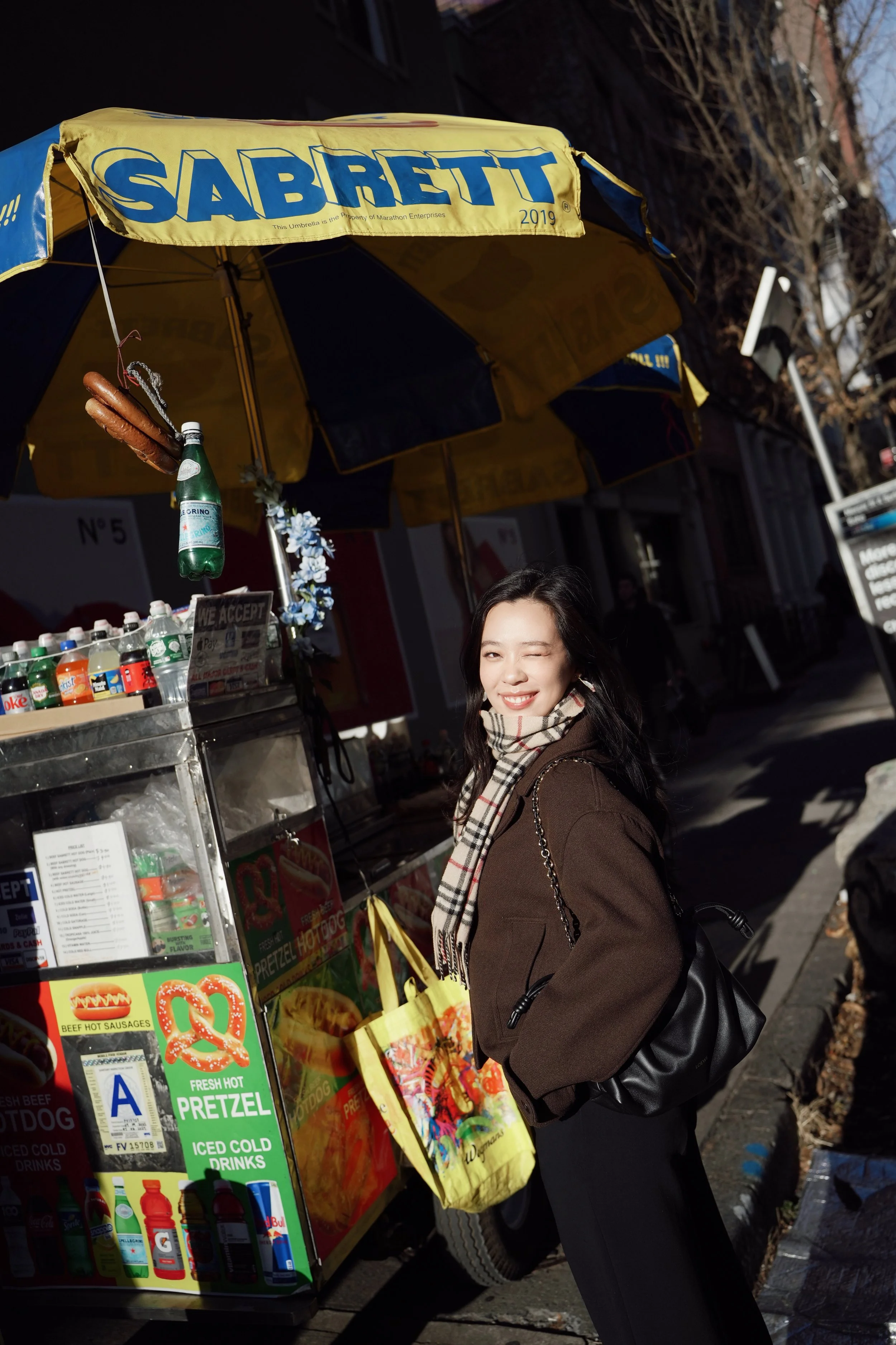 A woman standing in front of a hot dog and pretzel stand on a city street, wearing a brown coat and a plaid scarf, smiling and winking.