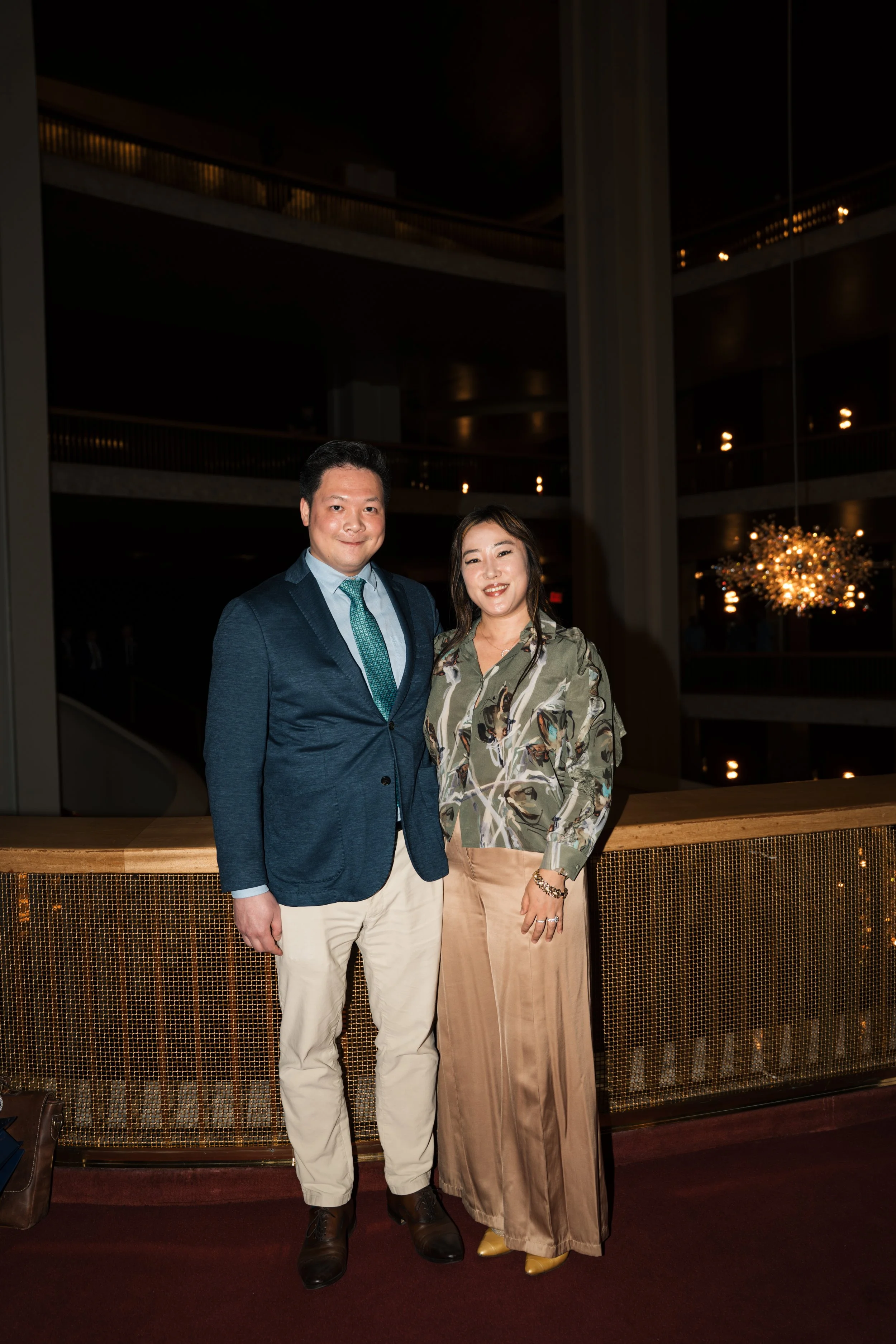 A man in a navy blazer, light blue shirt, and beige pants standing next to a woman in a satin beige skirt, patterned blouse, and yellow shoes. They are posing in a warmly lit, elegant interior with multiple levels and a chandelier in the background.