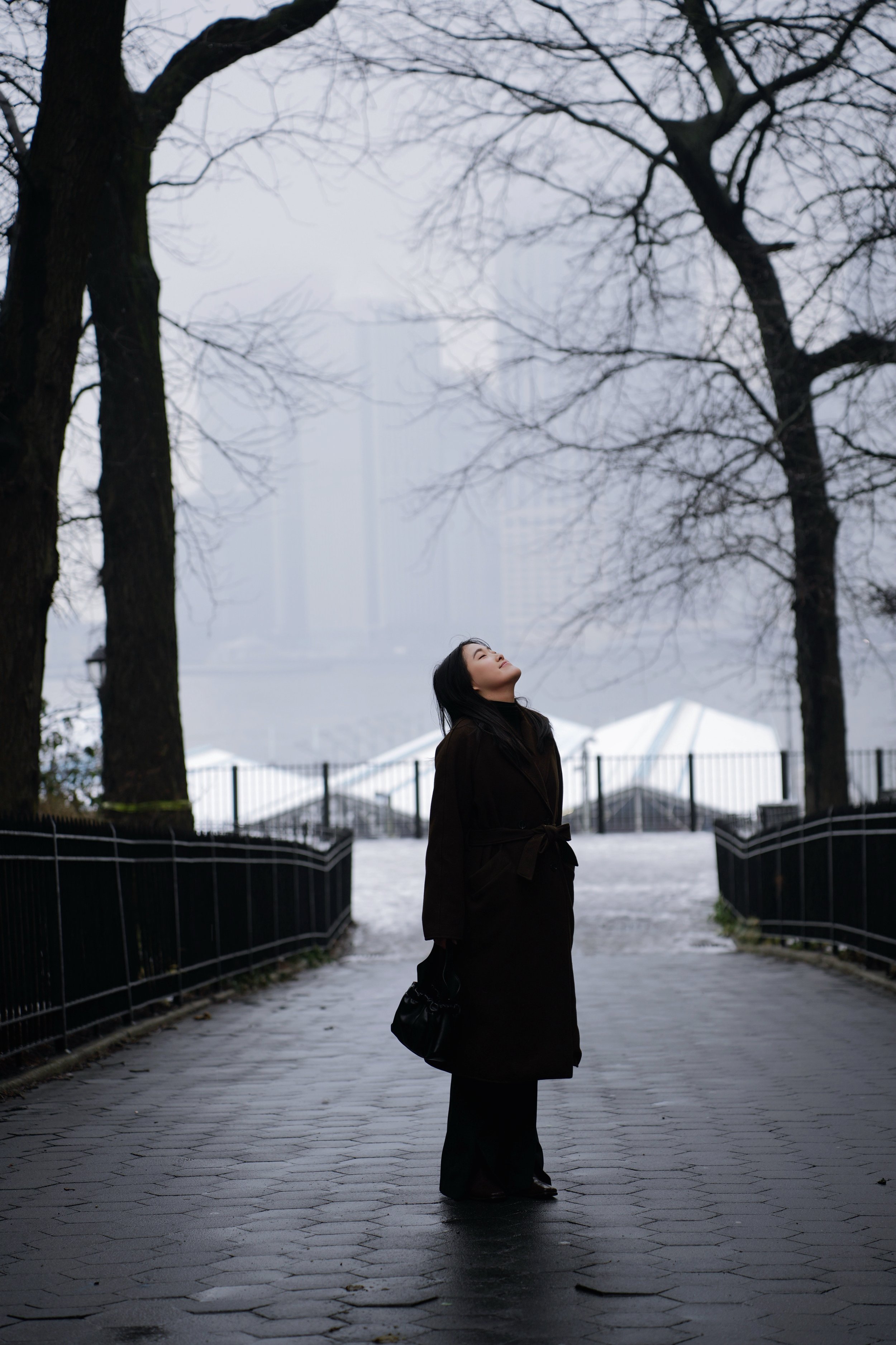 A woman in a dark coat standing outdoors on a cloudy day with her eyes closed and facing upward, surrounded by leafless trees and a snowy ground.