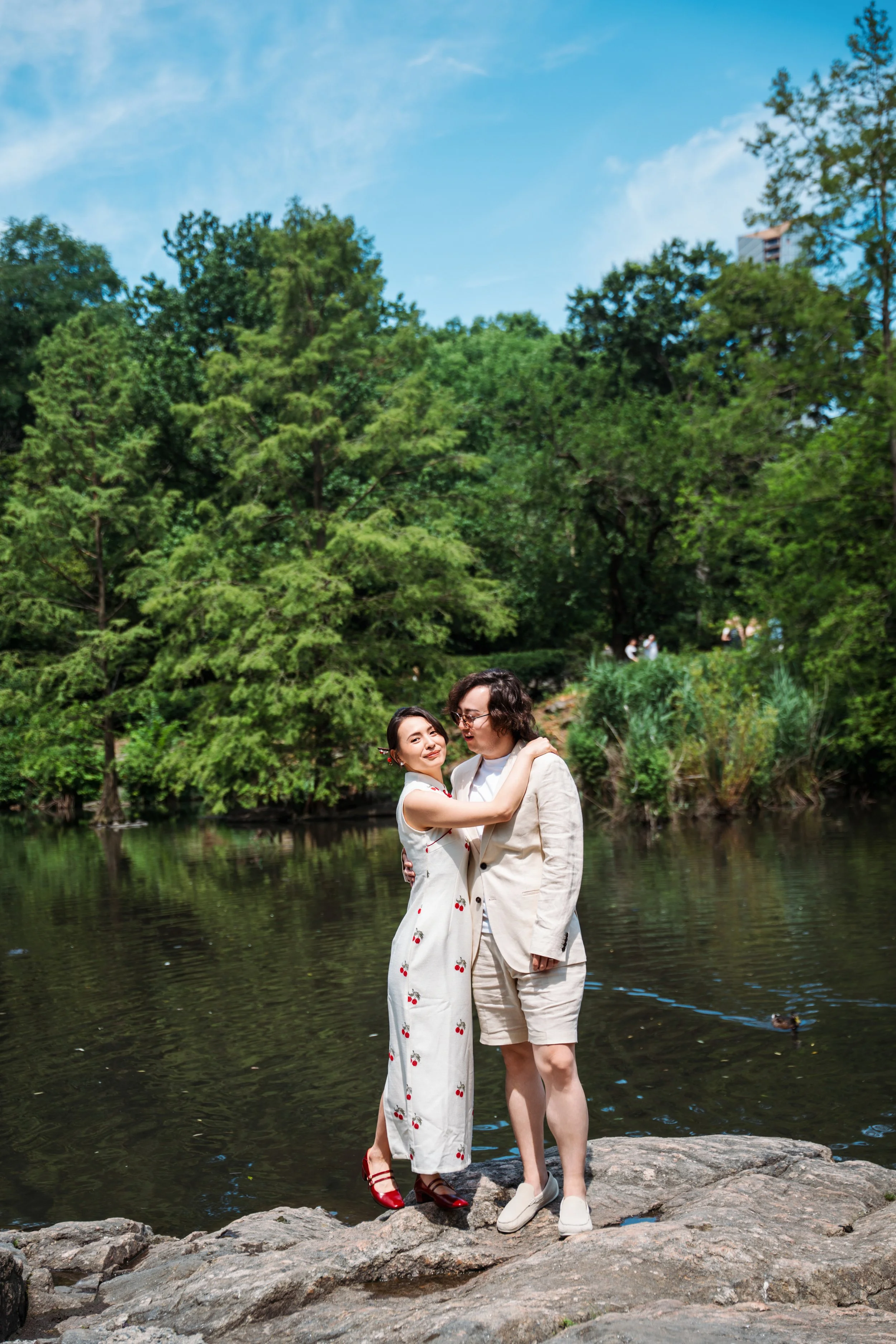 A couple standing on rocks by a river in a park with lush green trees and a blue sky in the background, embracing and smiling.