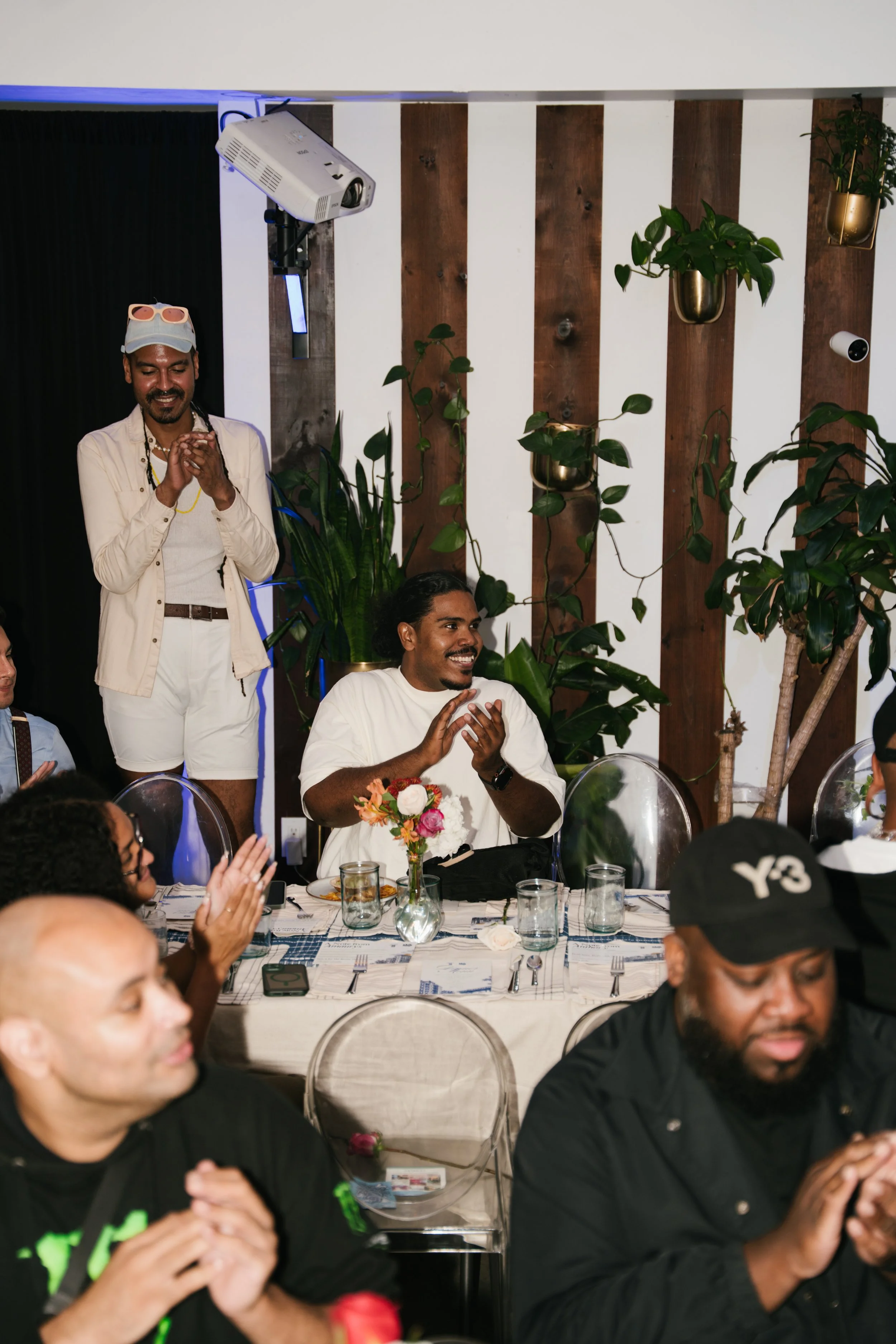 People gathered around a dining table at a celebration or event, some clapping and smiling. There are plants and a projector in the background.