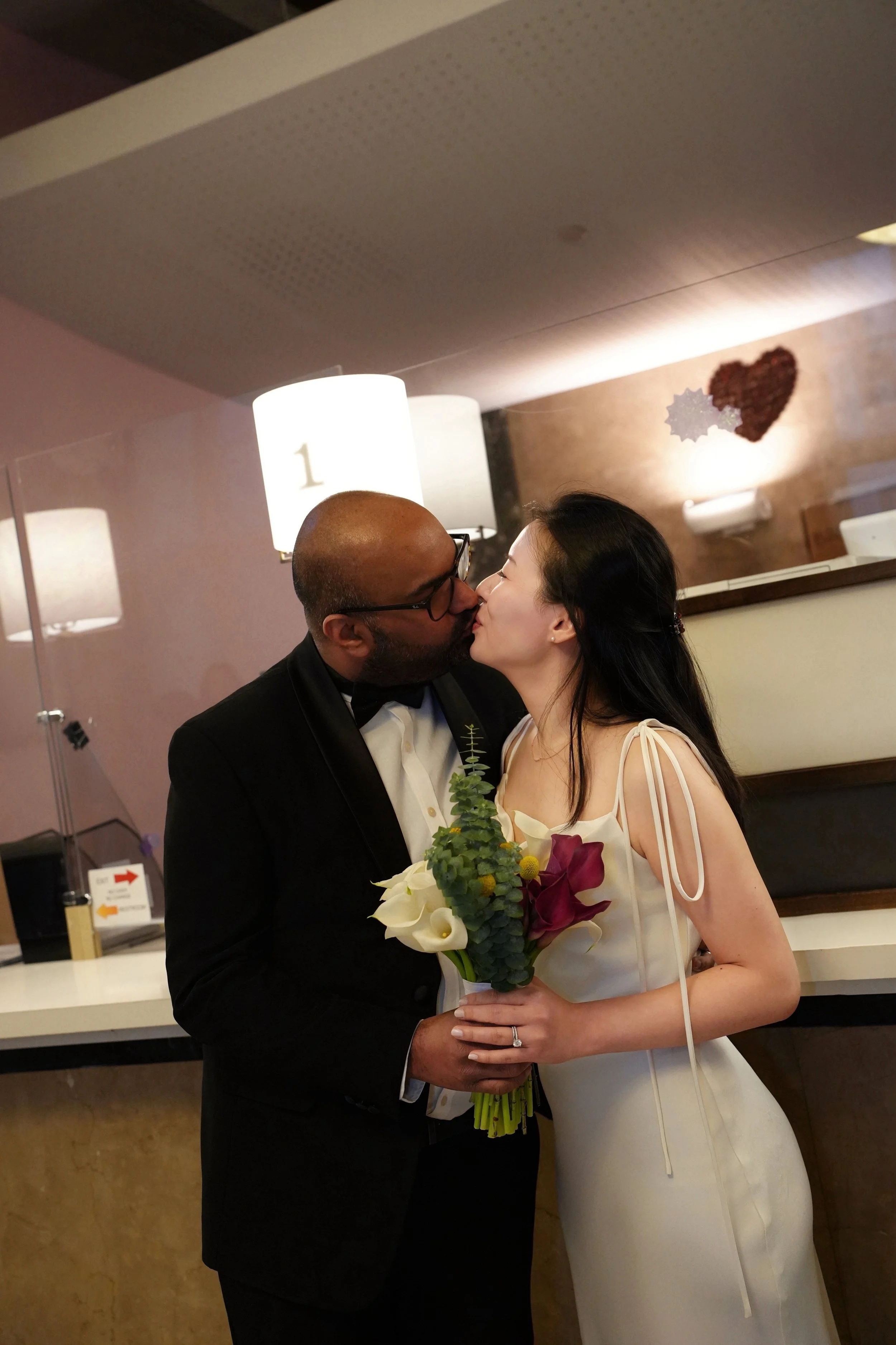 A man in a tuxedo and a woman in a white dress are kissing as she holds a bouquet of flowers.