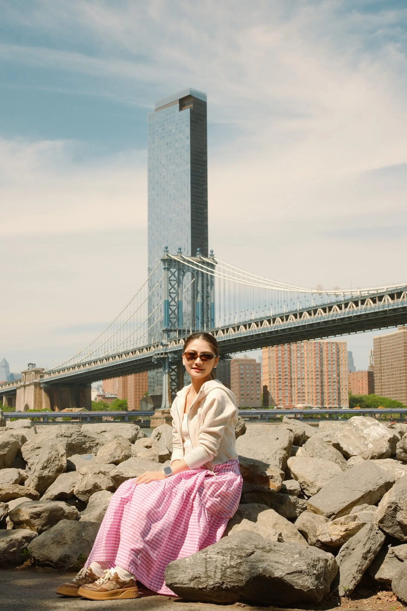 A woman wearing sunglasses, a beige hoodie, and a long pink checkered skirt is sitting on rocks by the water. Behind her, the Brooklyn Bridge and a tall modern skyscraper are visible under a partly cloudy sky in New York City.
