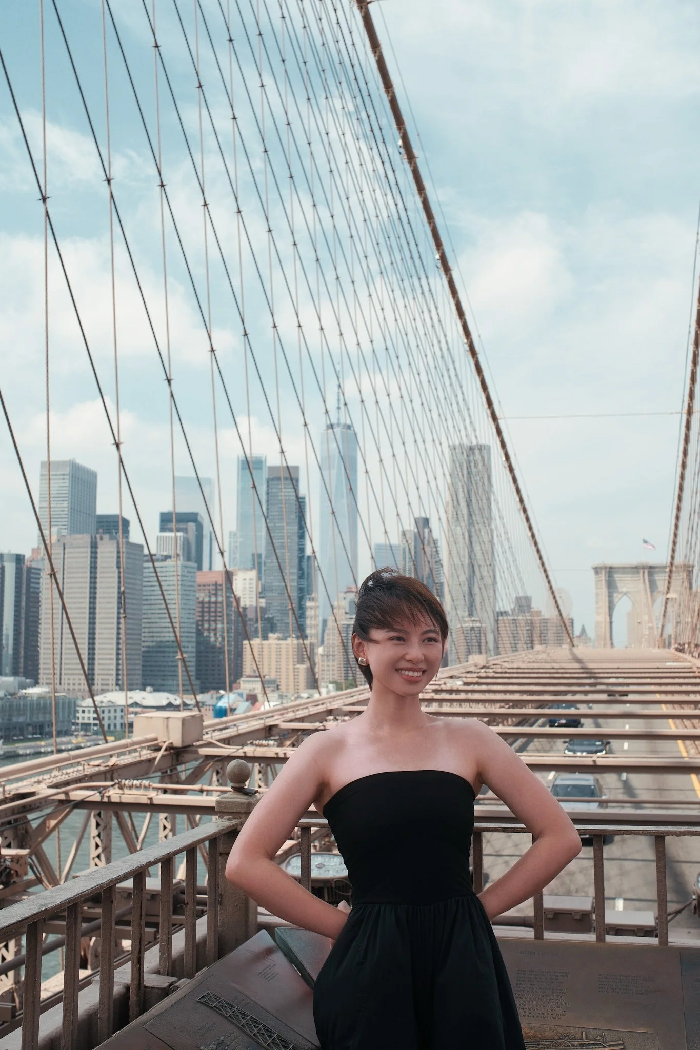 A young woman with short hair and earrings, wearing a strapless black dress, is standing on a bridge with her hands on her hips, smiling. The background features iconic NYC skyscrapers and the Brooklyn Bridge's cables and towers.