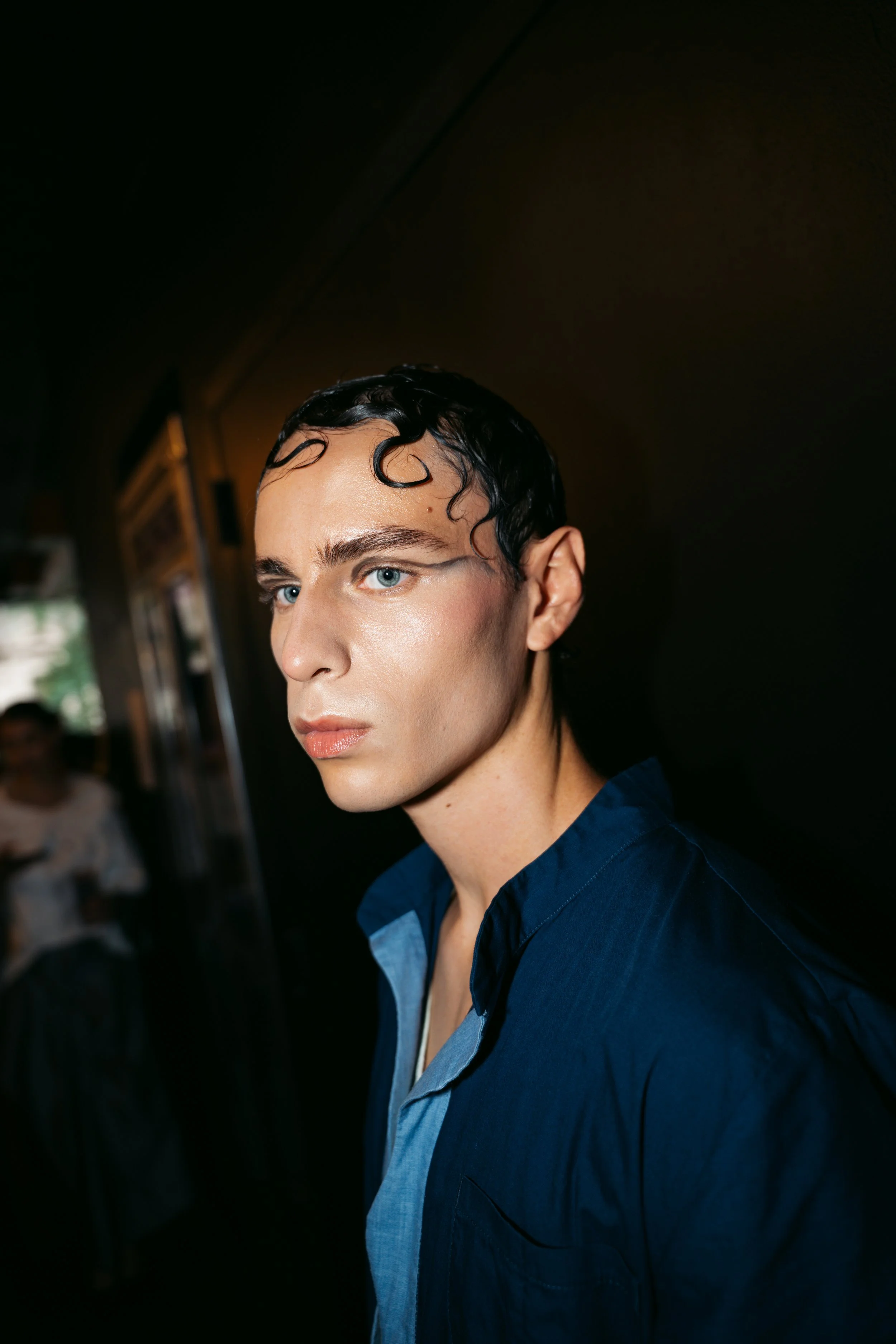 Close-up of a person with wet, black, slicked-back hair, wearing a blue shirt, standing indoors with a dark background.