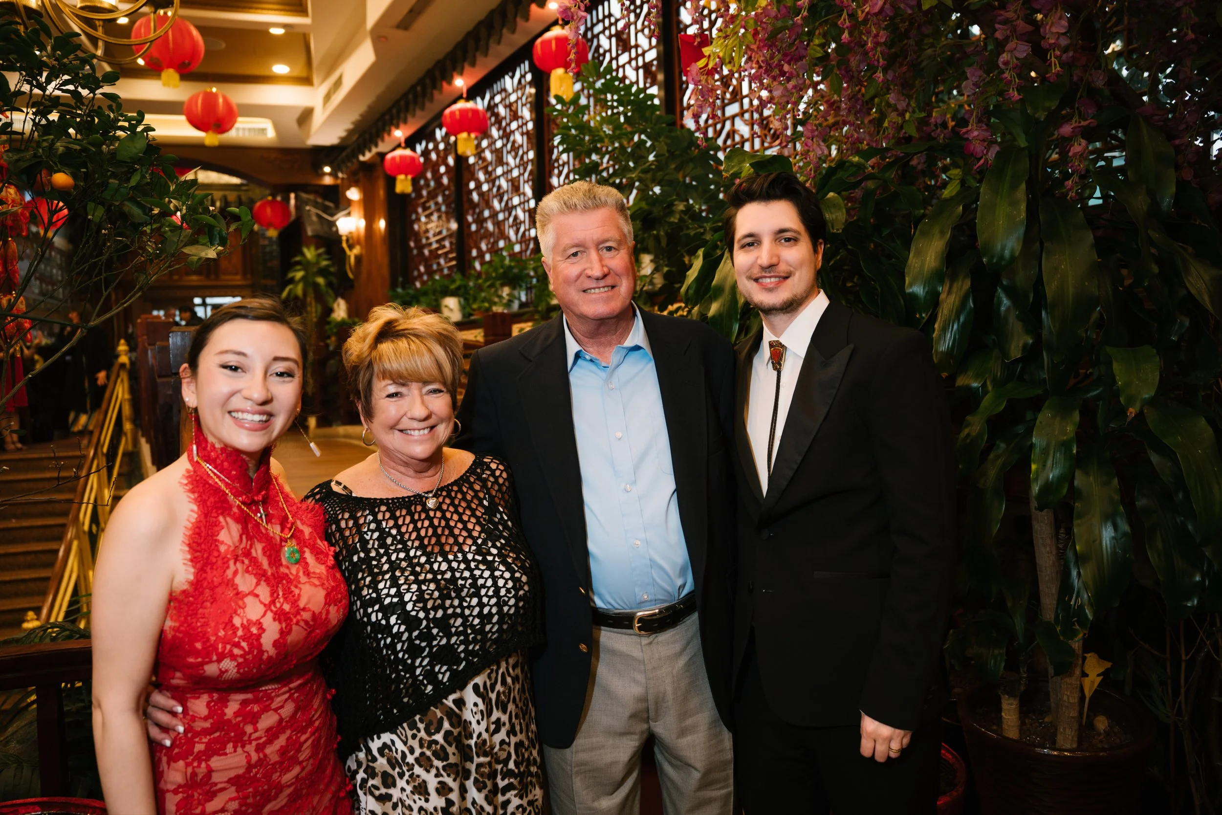 A group of five smiling people standing together in a decorated restaurant with red lanterns and lush green plants, celebrating an Asian cultural event.