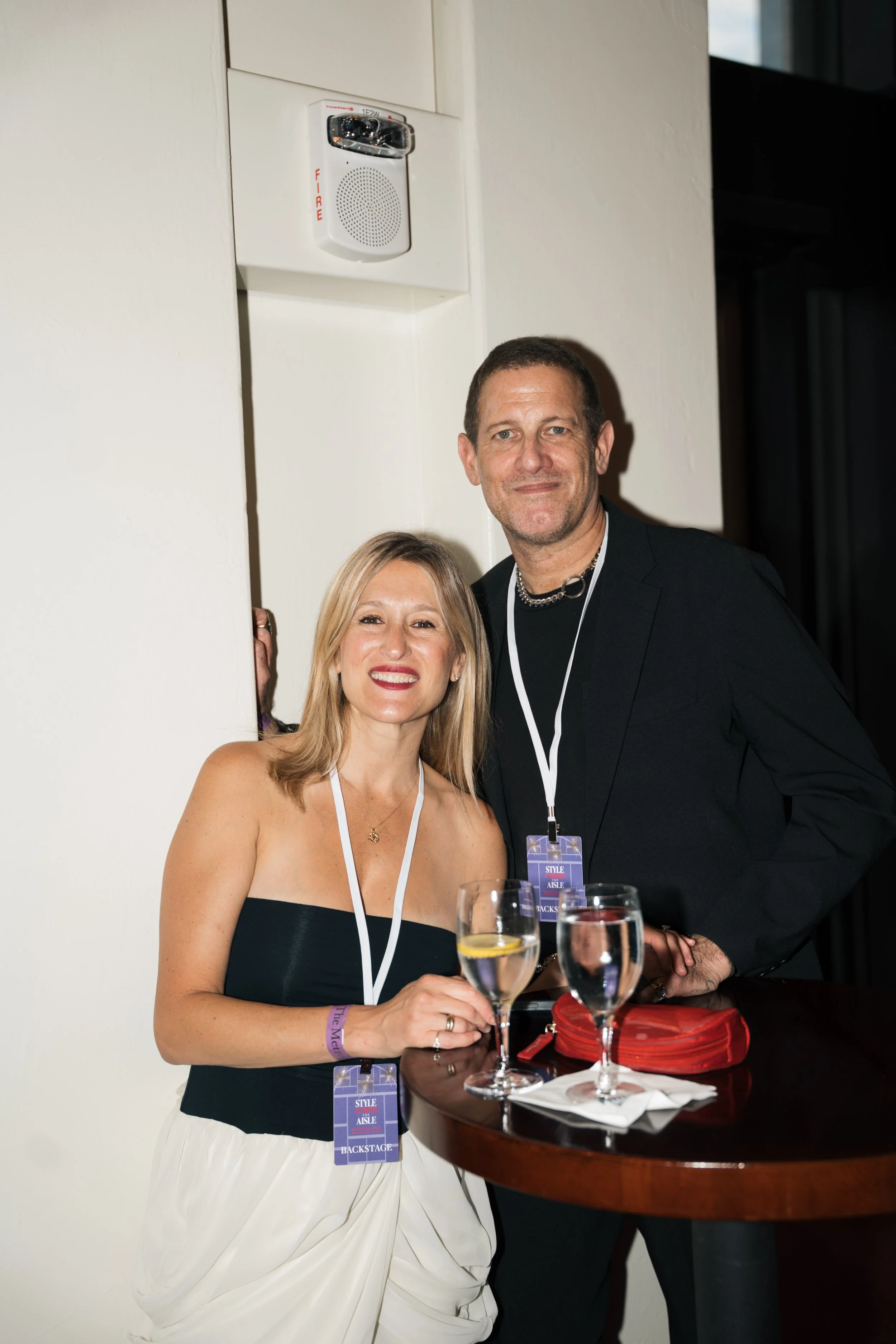 A woman in a strapless black dress with a white skirt and man in a black suit and necklace standing together at a social event, with a table holding wine glasses and a red clutch in front of them, in a room with white walls and a fire alarm.