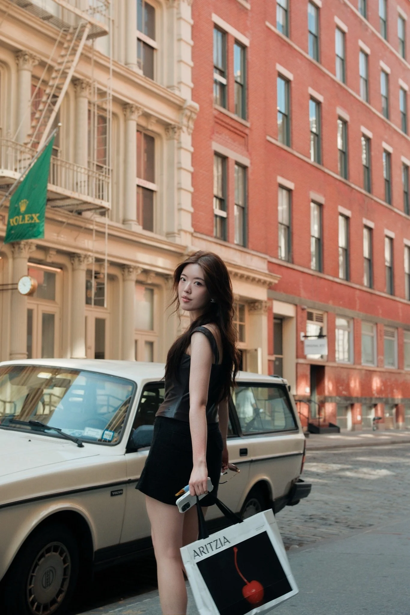 A young woman in a black dress carrying a shopping bag and a shopping guide, standing on a city street next to a vintage beige car, with brick buildings in the background.