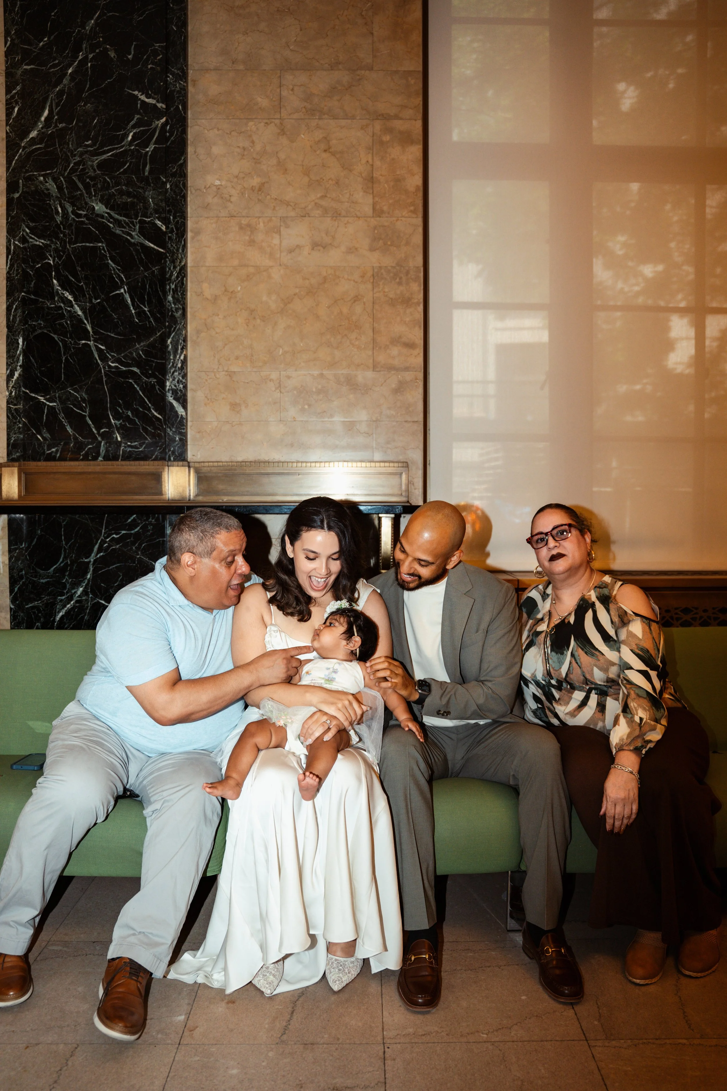 A group of six people, including a woman in a white dress, sitting on a green sofa in an indoor setting with marble walls and large windows. They are smiling and looking at an infant girl in the woman's lap, who is wearing a white dress and looking b