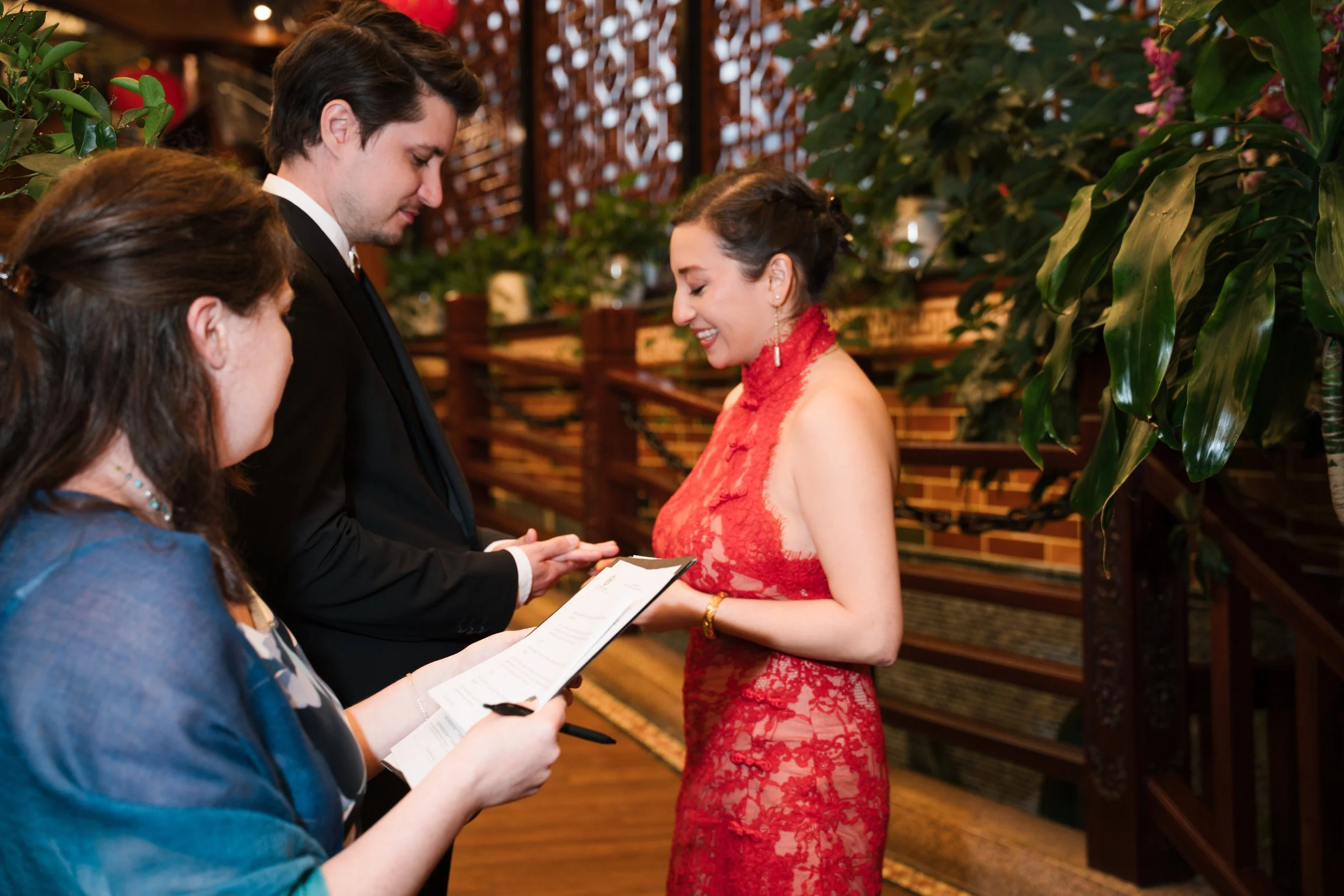 A wedding ceremony with a woman in a red lace dress and an officiant reading vows, with a man in a black tuxedo in front of her, and a woman in a blue dress holding a paper, in a warmly lit indoor venue with lush greenery.
