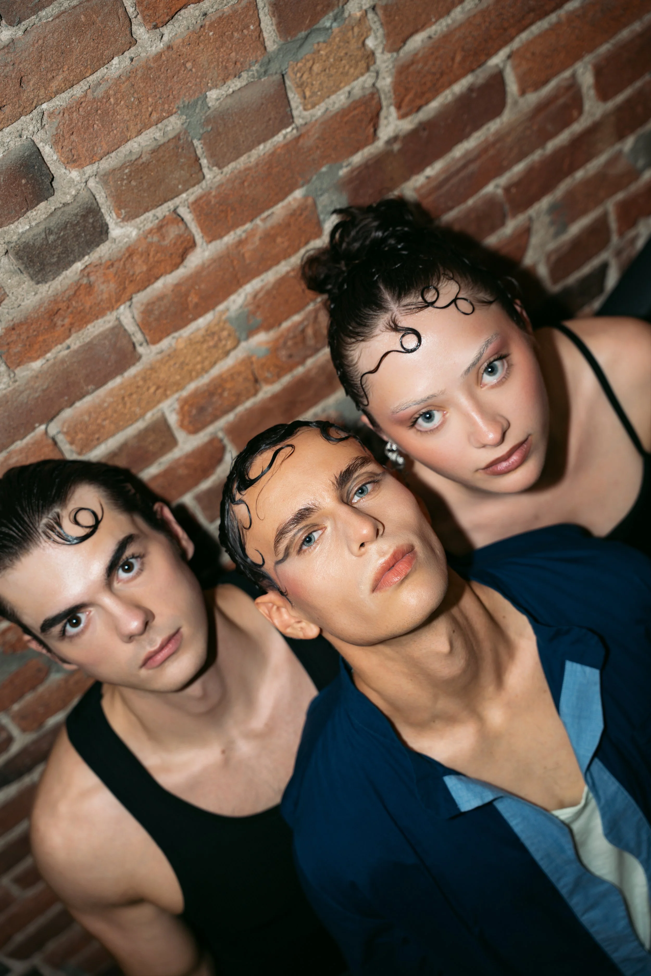 Close-up of five young women with curly hair and blue eyes, leaning against a brick wall, with distinct makeup and casual clothing.