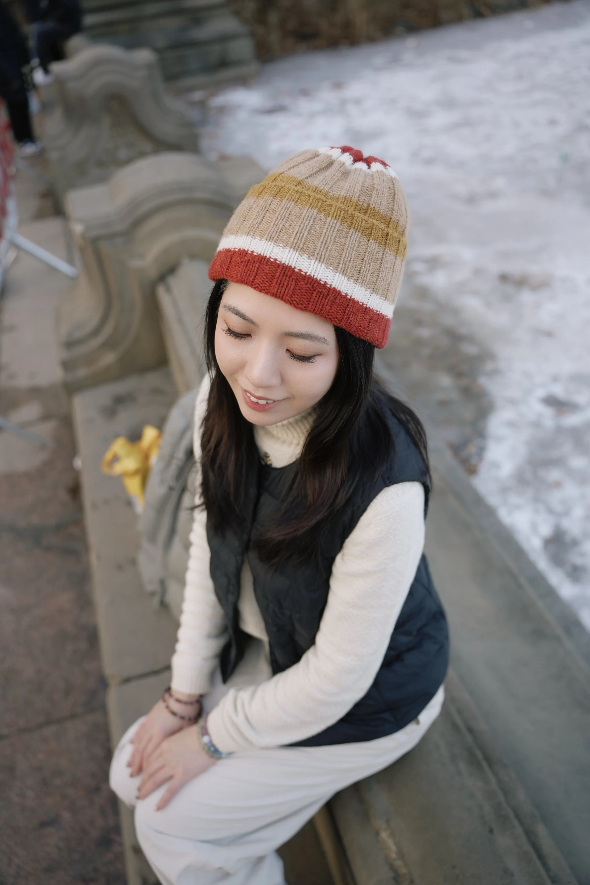 A young woman sitting on a bench outdoors, wearing a multicolored knit beanie, a cream sweater, and a black vest, with a snowy background.