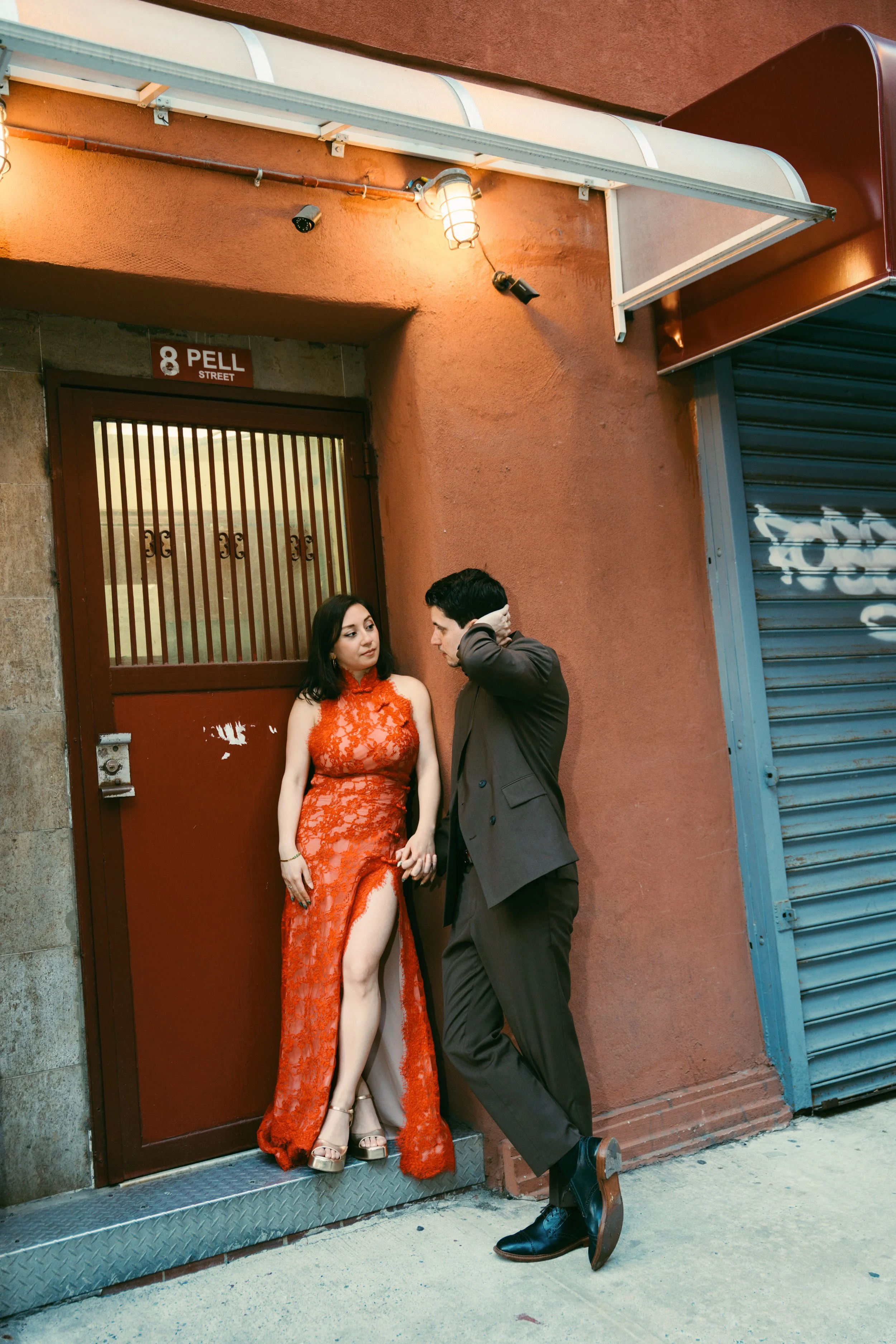 A man and woman standing on a city street near a building with a red wall. The woman is wearing an elegant, long, red lace dress with a high slit, and the man is in a dark suit. They are looking at each other, with the woman holding her dress and the