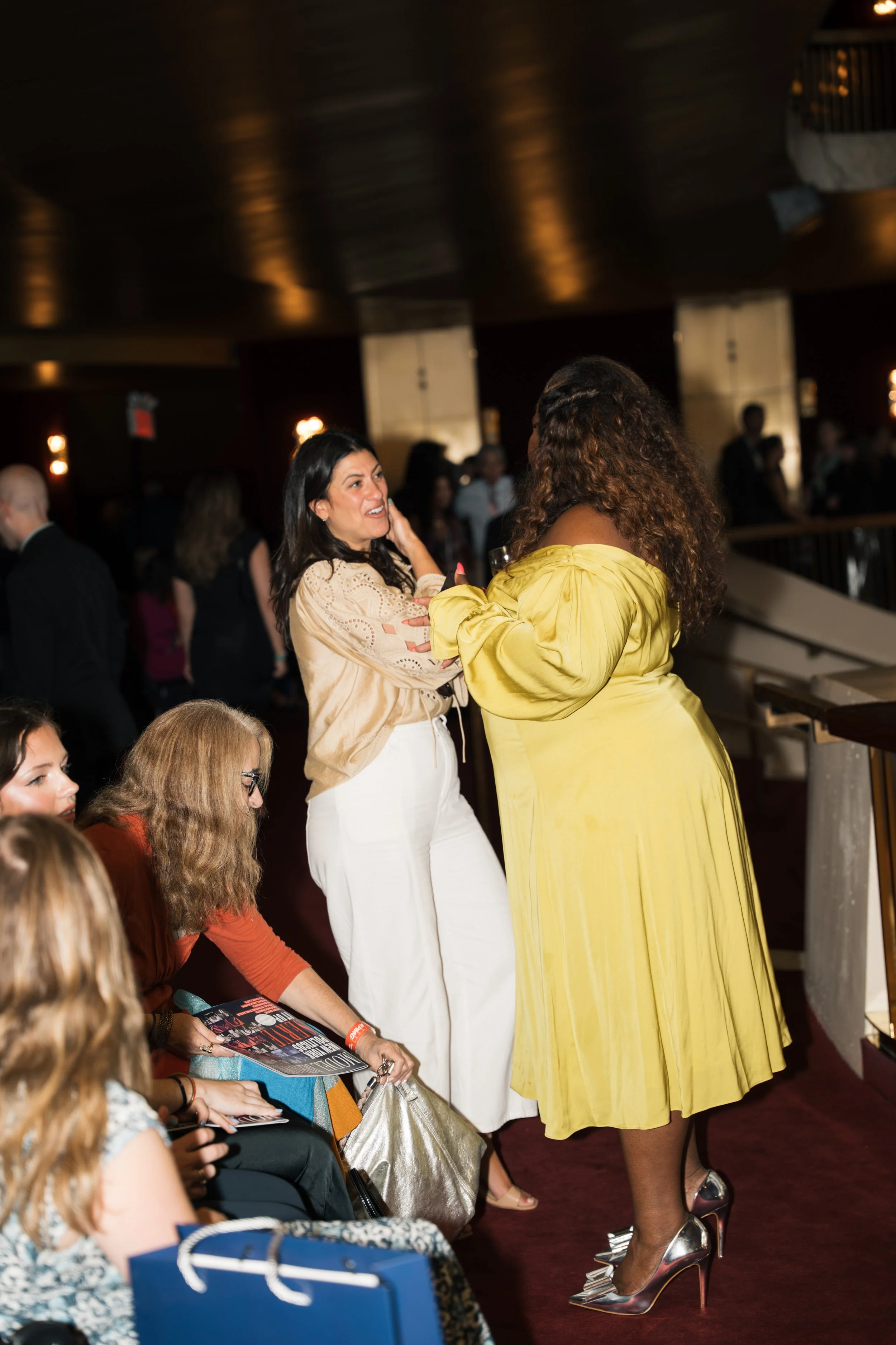 Two women having a conversation at an event, with several seated people observing, one woman in a cream-colored blouse and white pants, the other in a bright yellow dress, both smiling. The setting appears to be a formal or social gathering in an ind