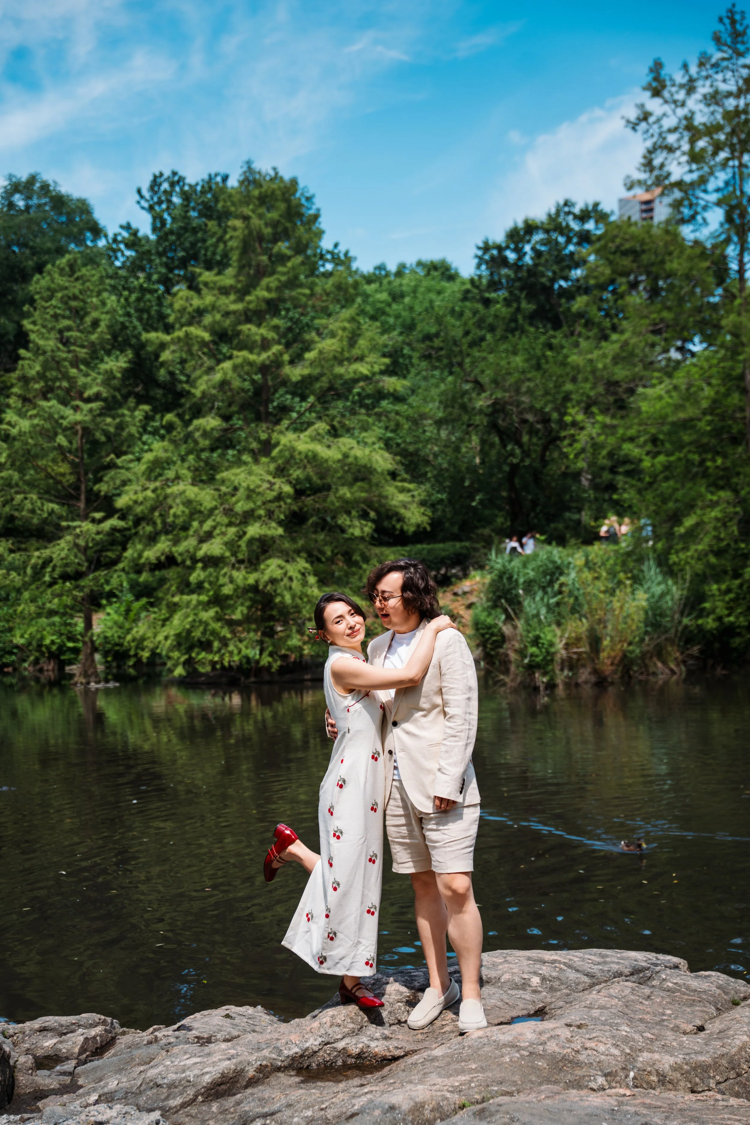 A couple stands on rocks near a river, embracing, with trees and a blue sky in the background.