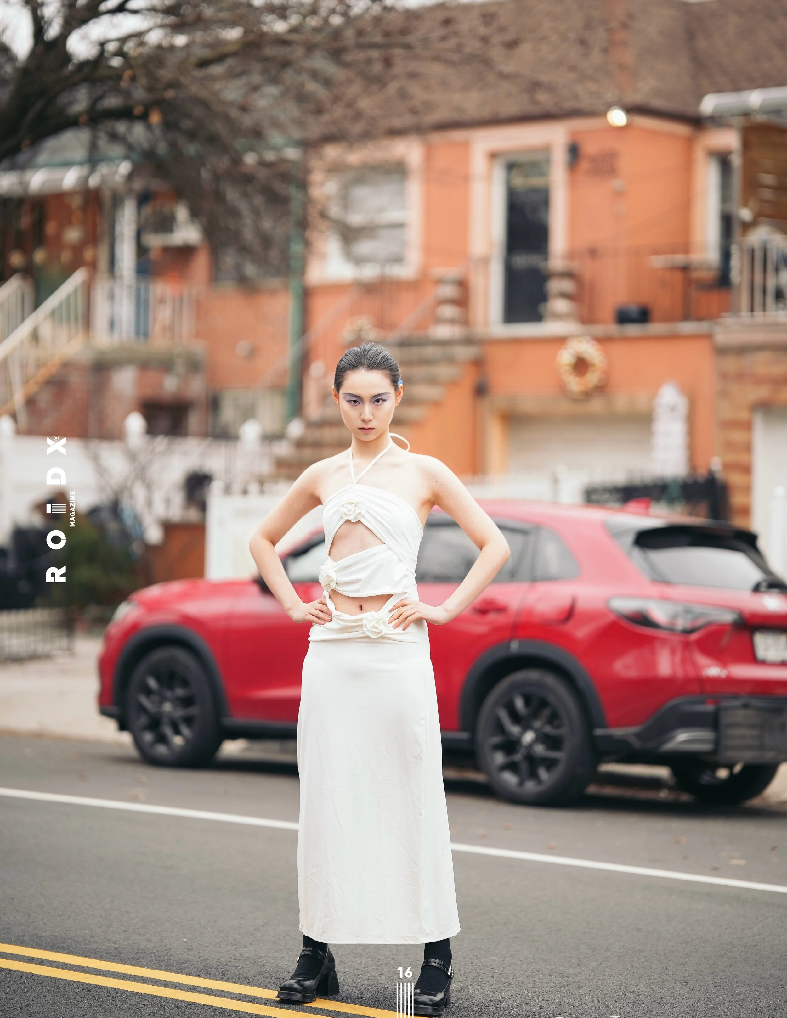 A young woman standing on a street with her hands on her hips, wearing a white strapless dress, black boots, and dark hair pulled back. Behind her, there is a red car and a brick house with a wreath decoration, trees, and other houses in the background.