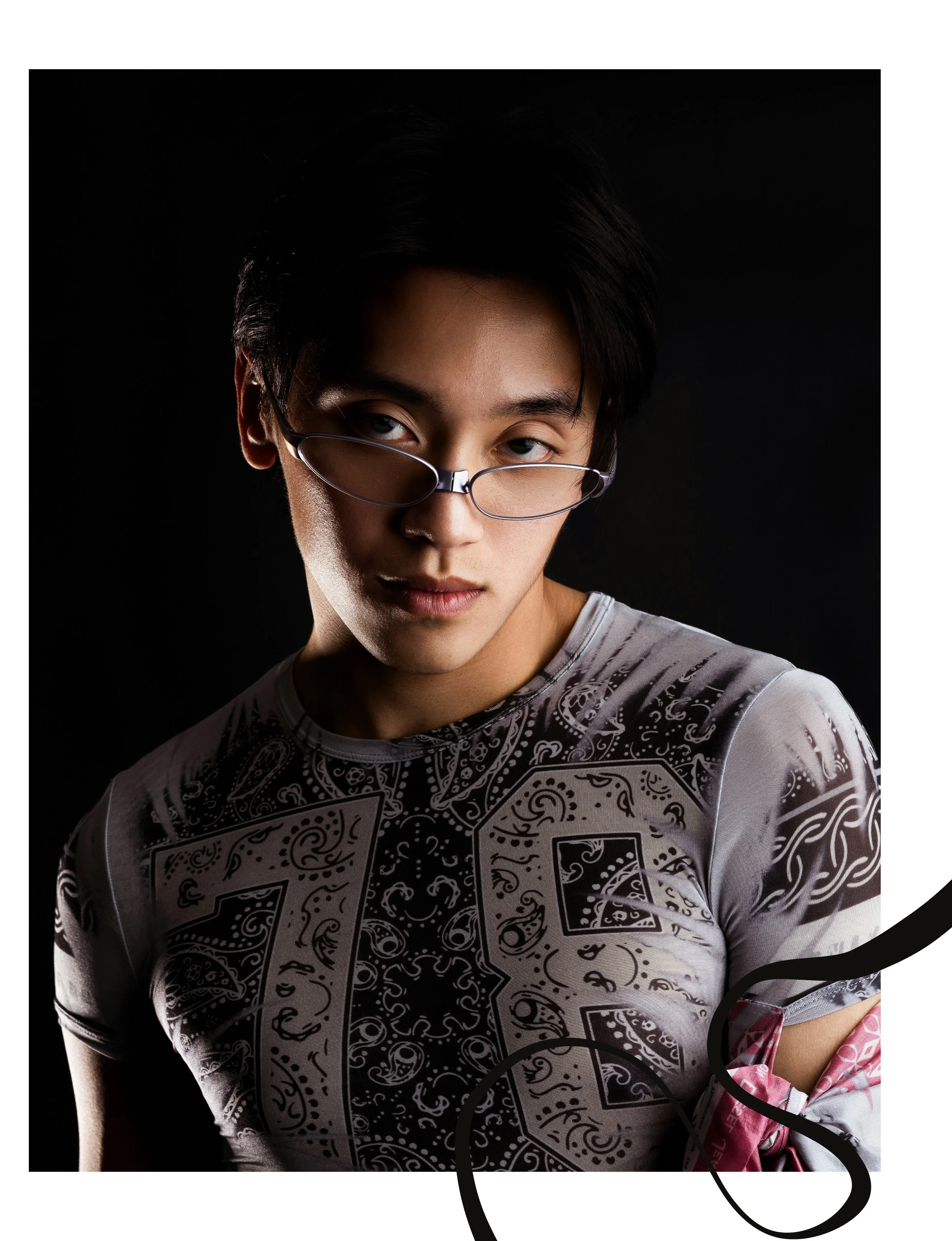 A young man with black hair and glasses looking directly at the camera, wearing a patterned shirt, against a black background.