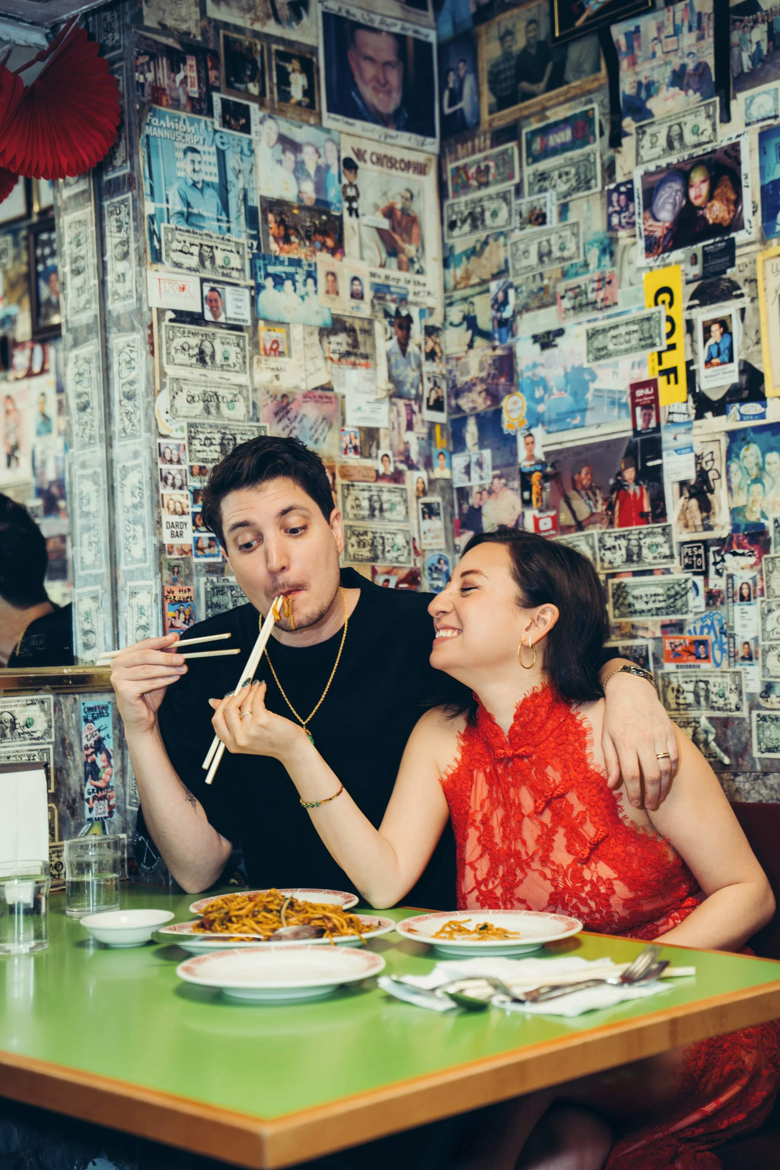 A young man with dark hair wearing a black shirt and gold necklace eats with chopsticks while a woman with long dark hair and a red lace dress smiles and holds his hand at a restaurant table. The table has various plates of food, including noodles, a