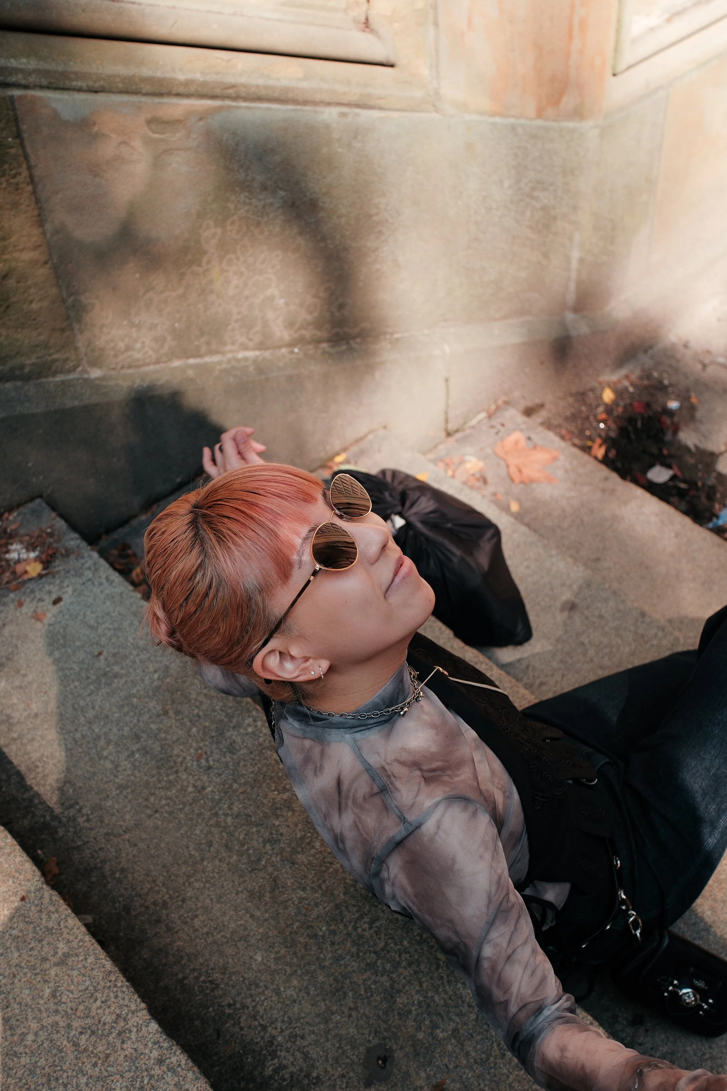 A person with orange hair and sunglasses sitting on the sidewalk, leaning against a stone wall with dirt and leaves around.