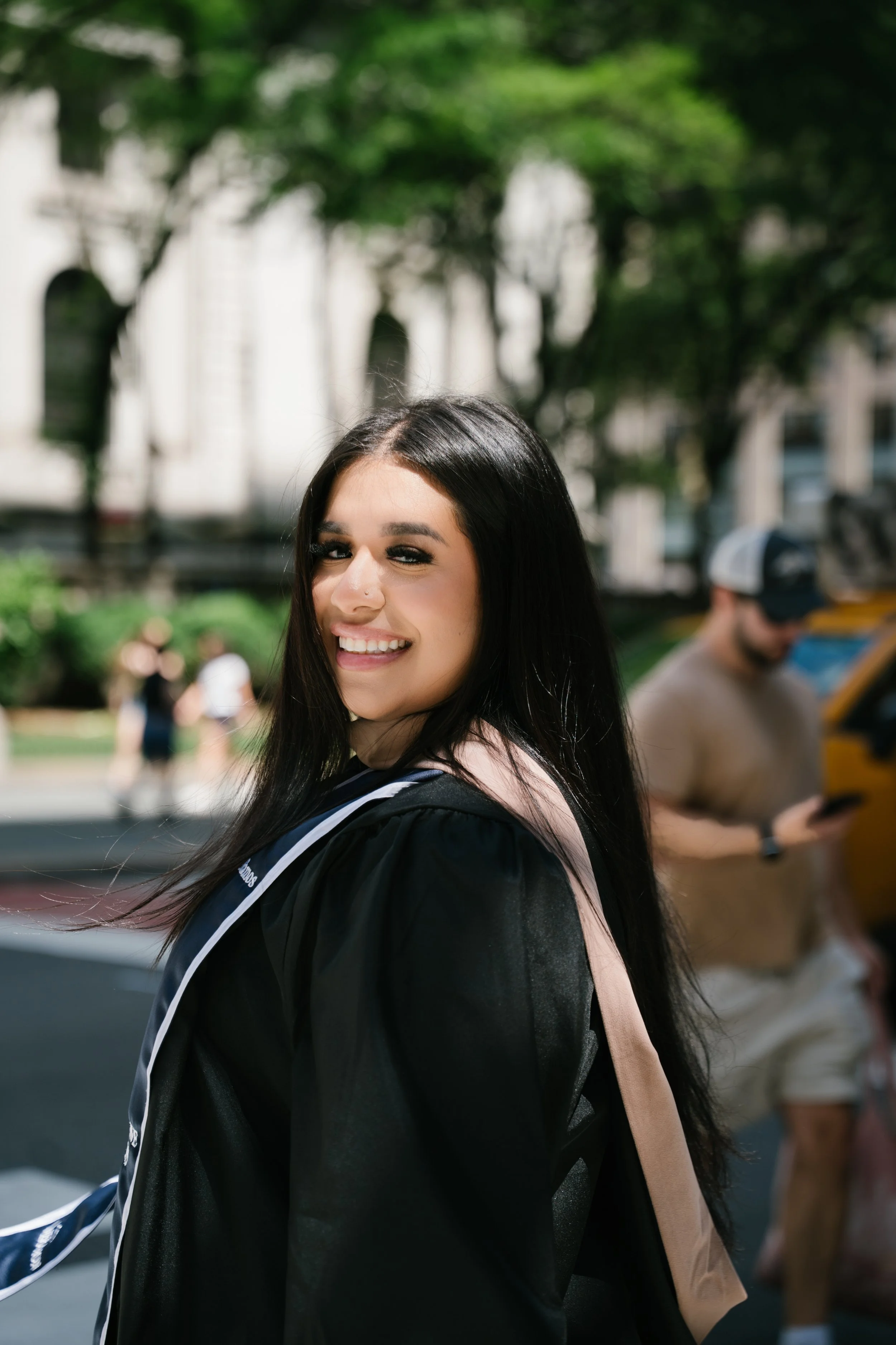 A young woman with long black hair smiling outdoors in the daytime, wearing a light pink top and a black jacket with a backpack. There are trees and a person looking at their phone in the background.