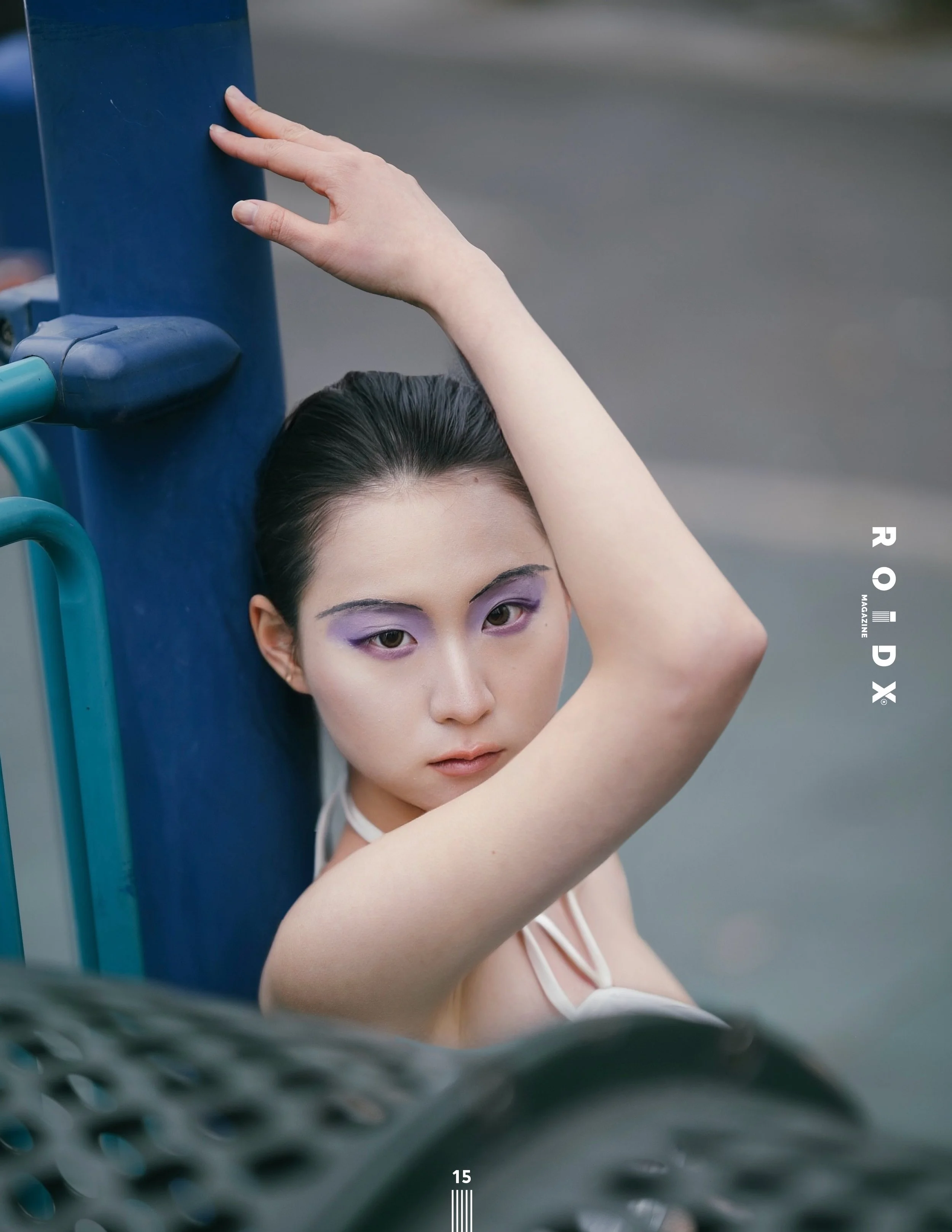 Close-up of a woman's face with purple eye makeup, hand resting on a blue parking meter, with a blurred background of pavement.