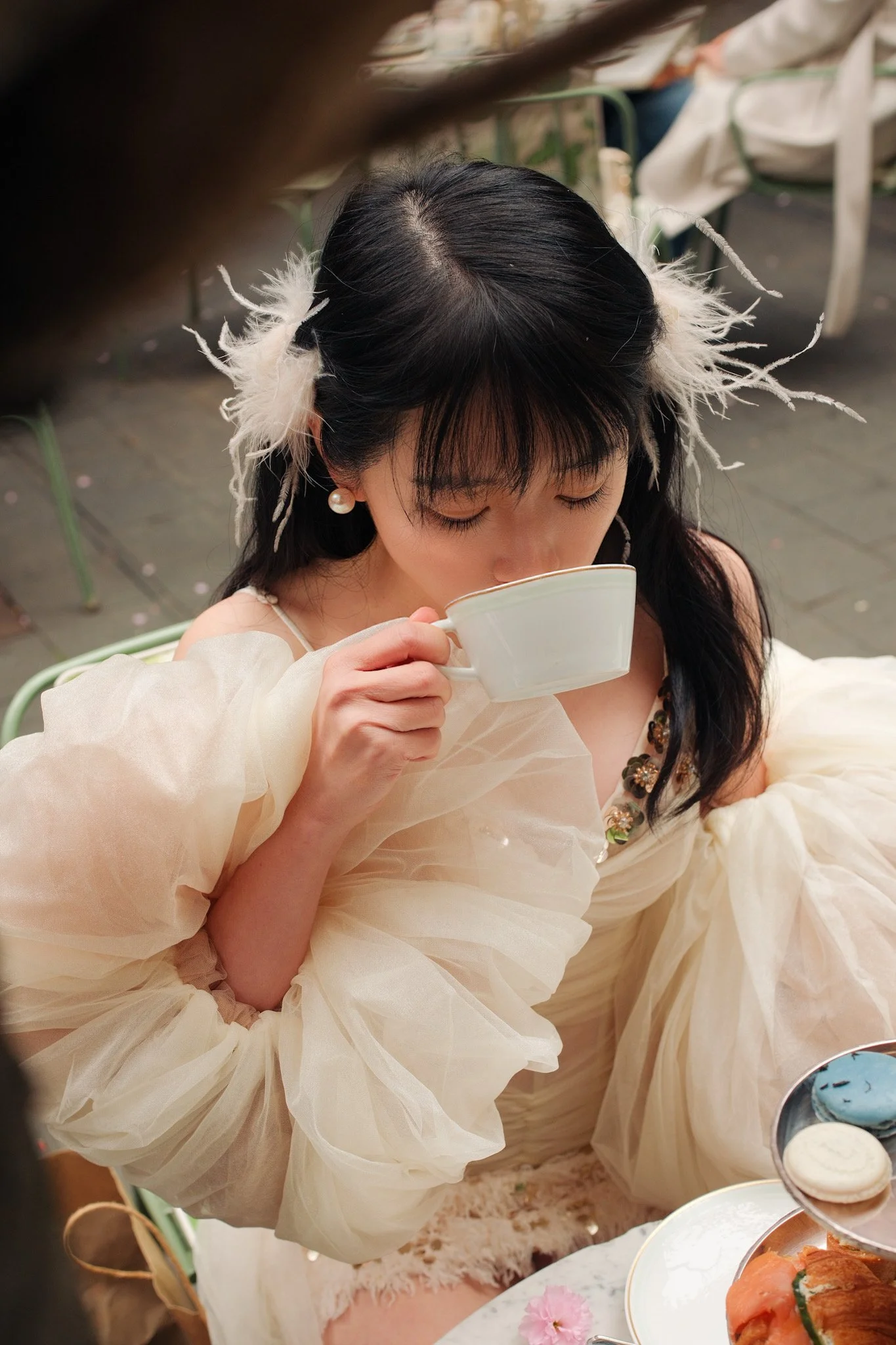 Young girl with dark hair, wearing pearl earrings and a light-colored, fluffy dress, is holding a teacup close to her face and appears to be drinking or smelling tea at a tea party with pastries and desserts on the table.