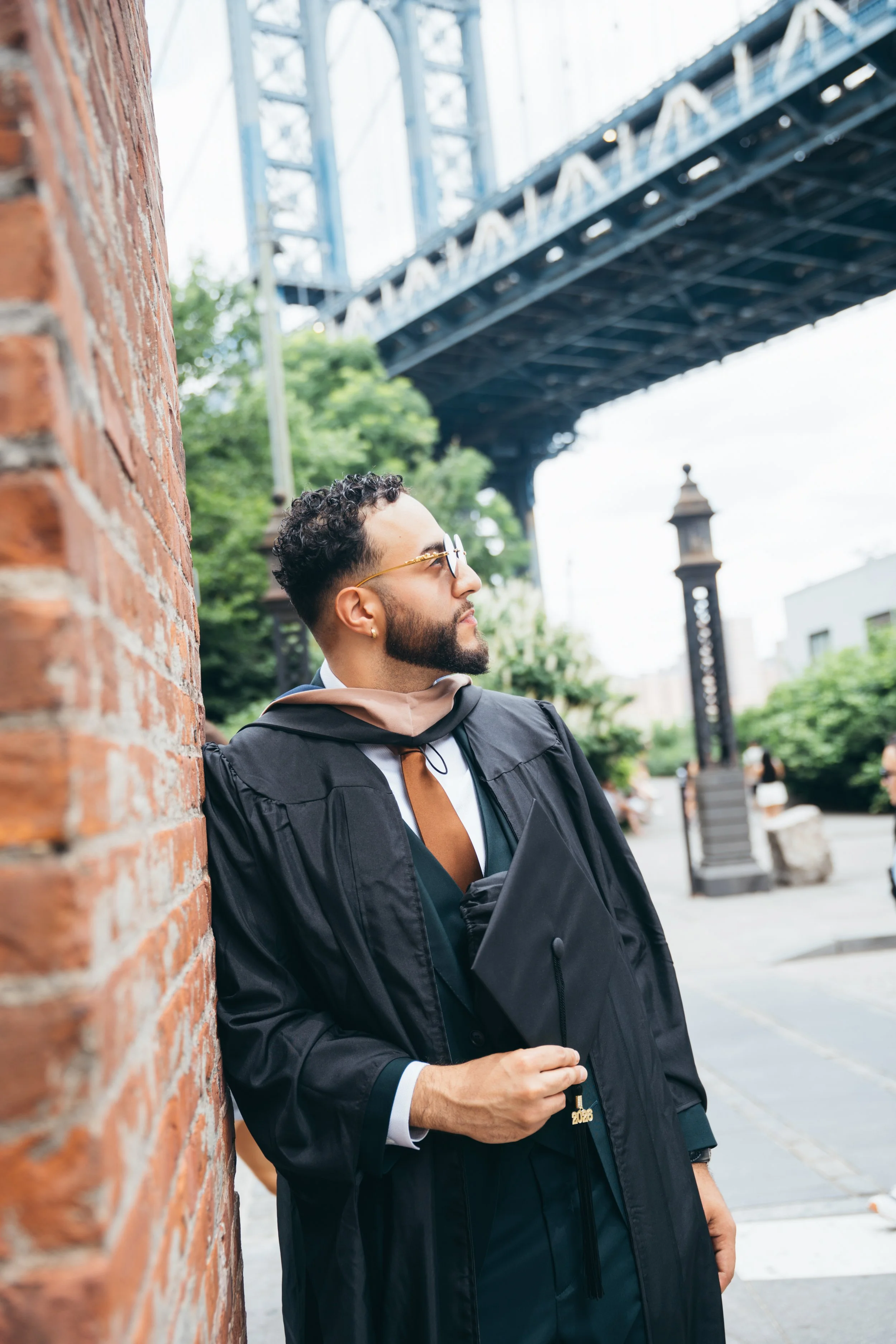 A young man with curly hair, beard, sunglasses, and earrings standing against a brick wall in an outdoor urban setting, wearing a graduation gown and holding a graduation cap that says "2026". There is a bridge above and trees in the background.