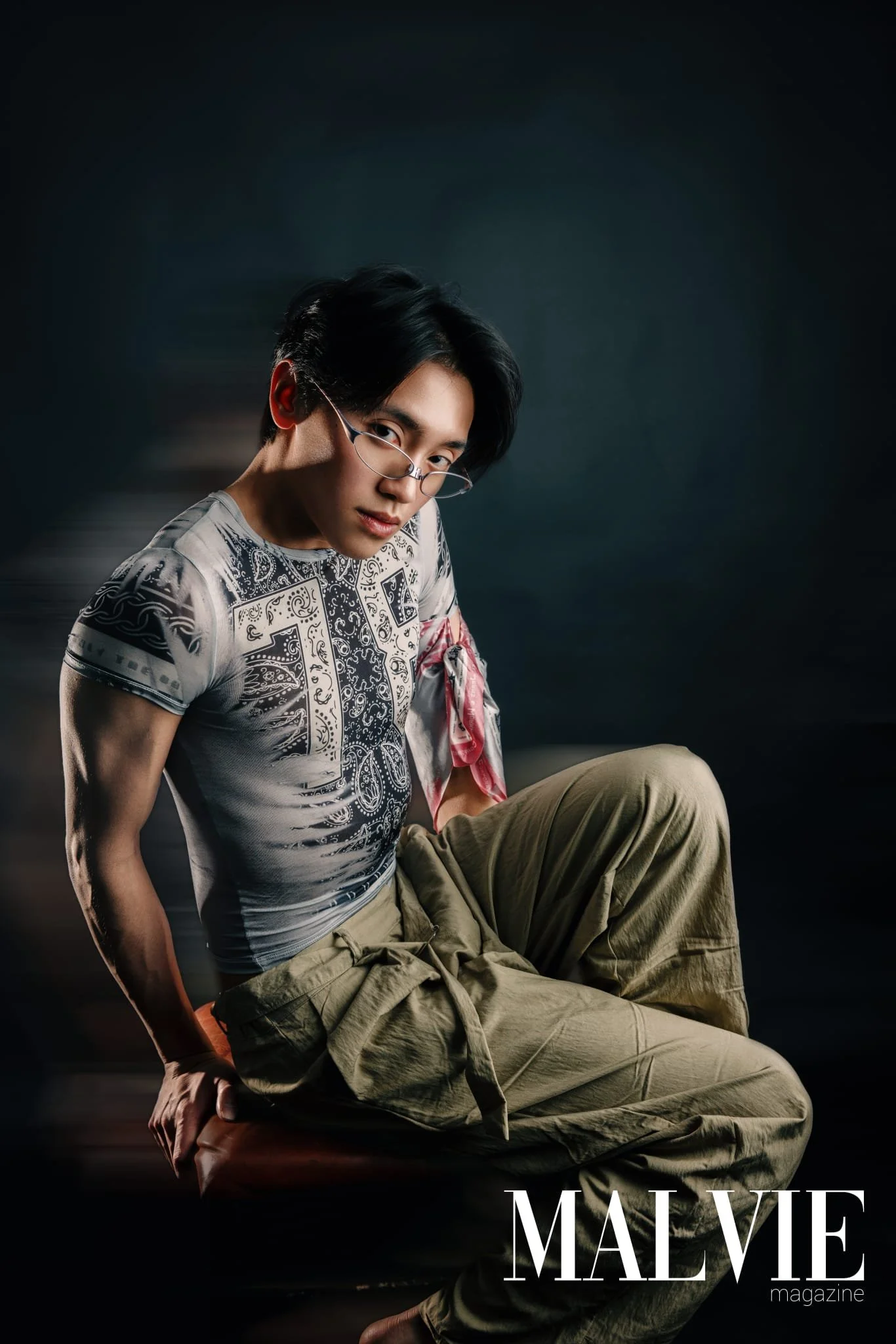A young man with black hair wearing glasses, a patterned T-shirt, and beige pants poses against a dark background for a magazine cover titled 'MALE'.