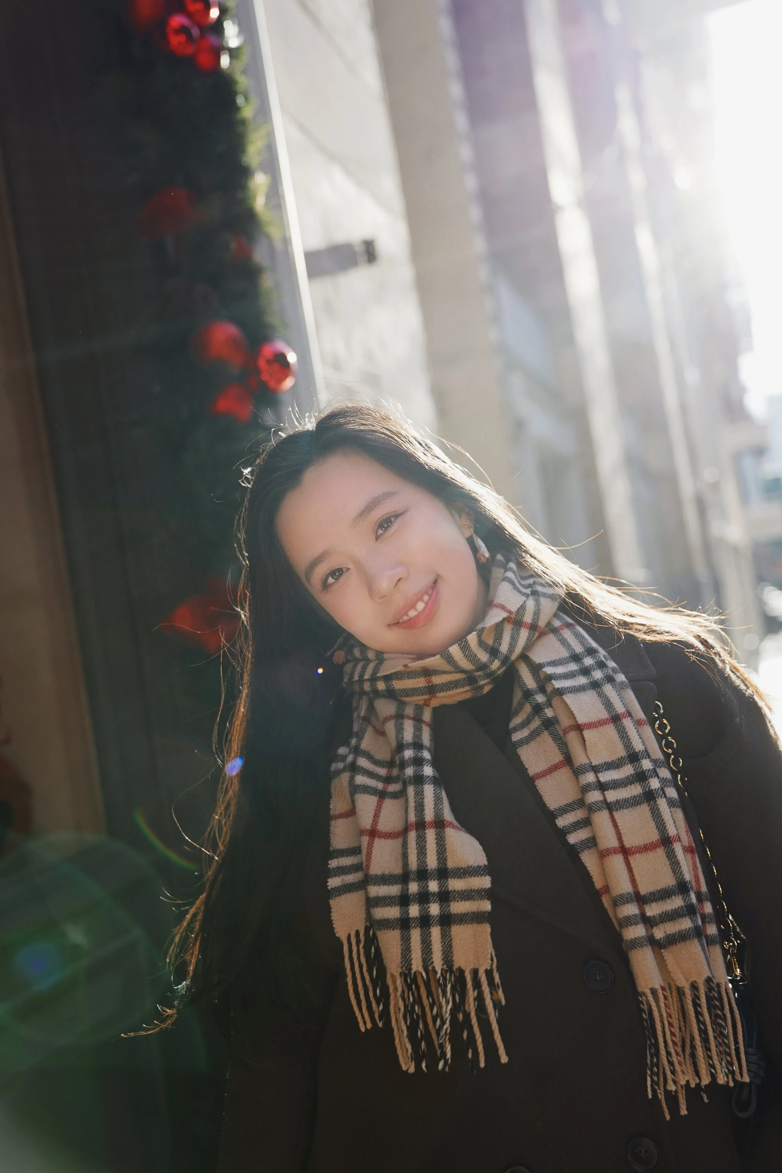 Young woman with long dark hair smiling, wearing a black coat and a beige plaid scarf, standing near a window with sunlight, with blurred red ornaments and greenery reflection visible.