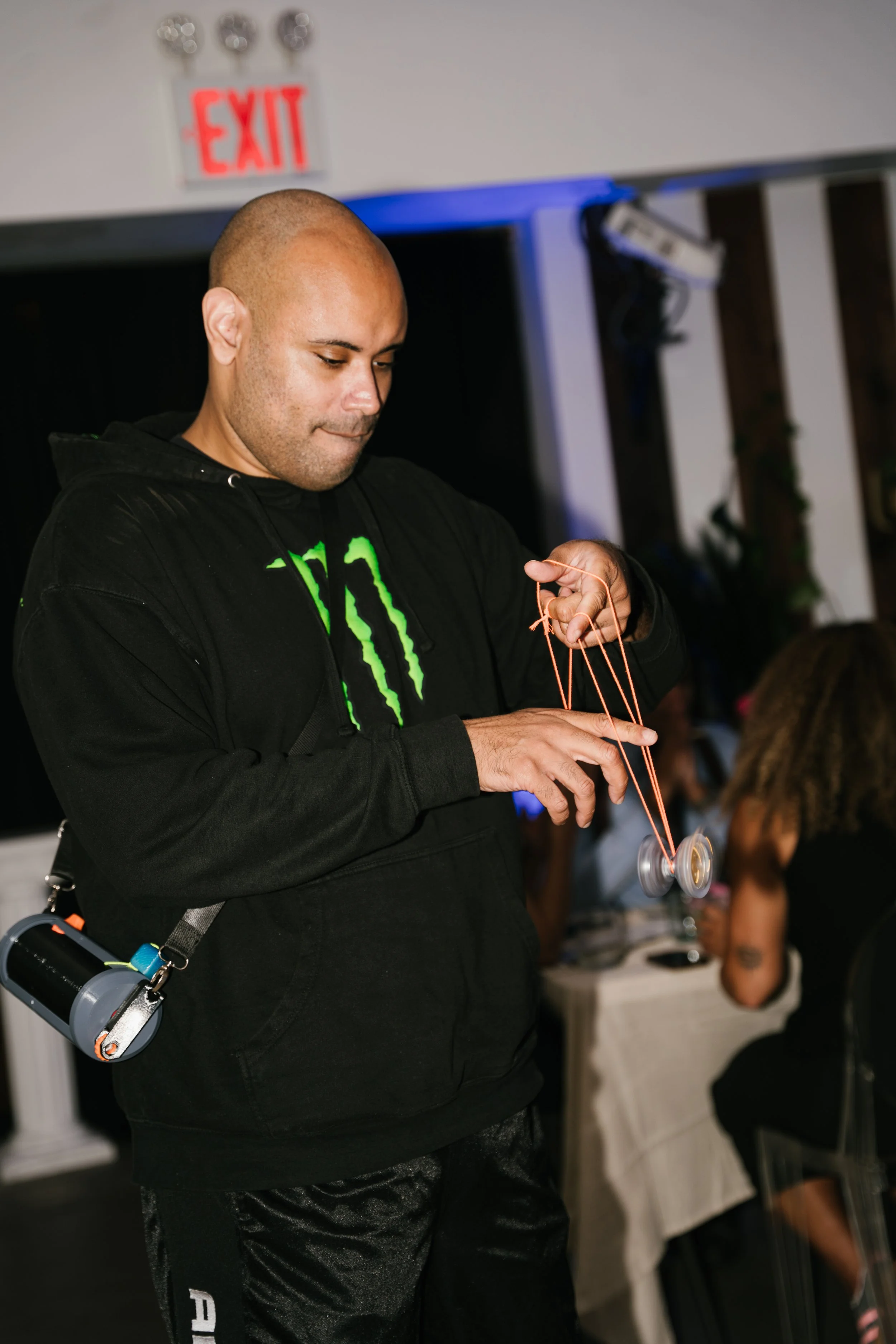A man with a shaved head wearing a black hoodie with a neon green Monster logo is using a yoyo at an indoor event. In the background, there's an illuminated exit sign and a woman sitting at a table.