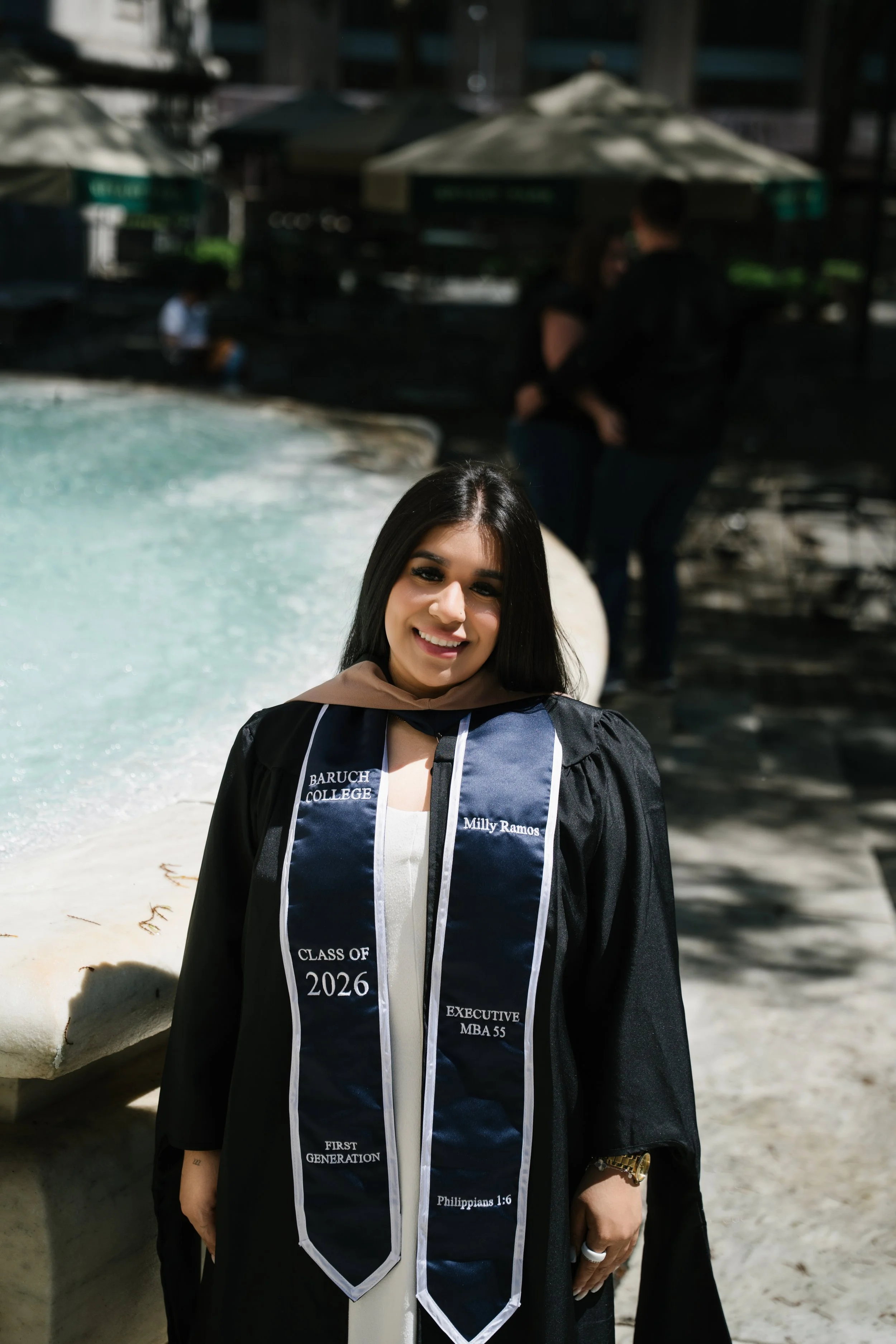 A young woman wearing a graduation gown and stole with academic and Biblical inscriptions, smiling outdoors on a sunny day with a fountain and people in the background.