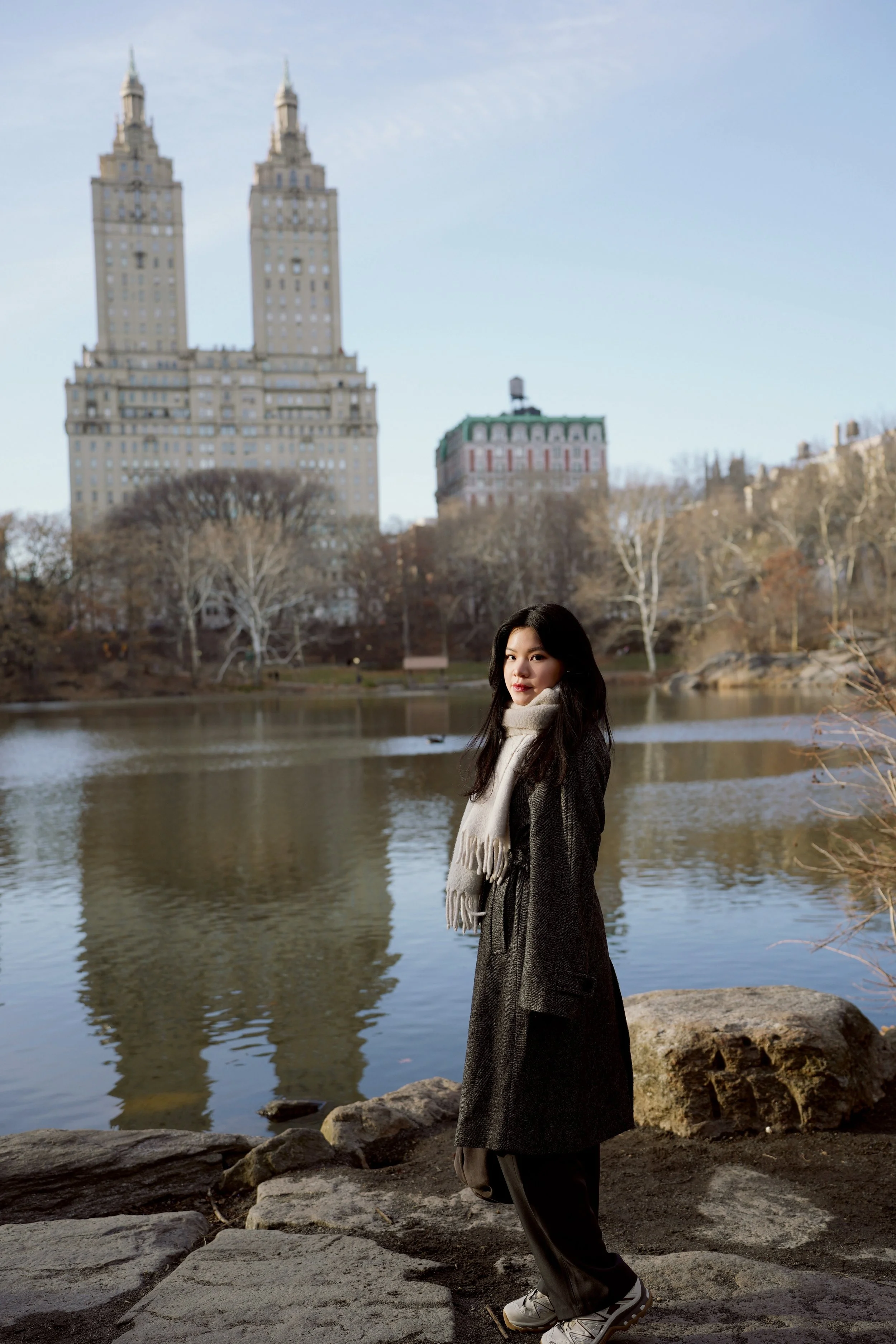 Young woman standing near a lake with trees and tall buildings in the background on a cloudy day