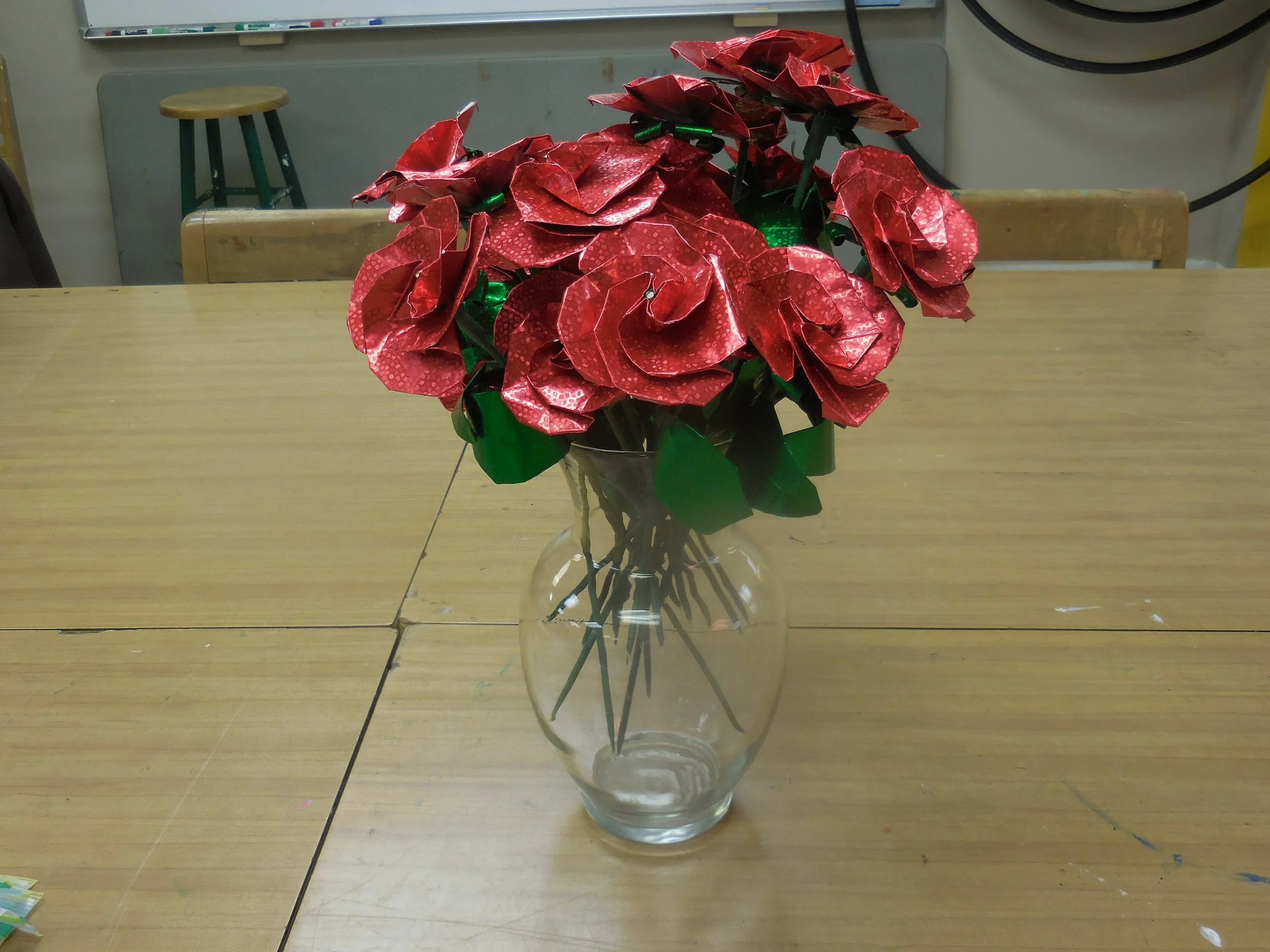 A glass vase with a bunch of red metallic artificial roses on a wooden table, with a classroom whiteboard and chairs in the background.