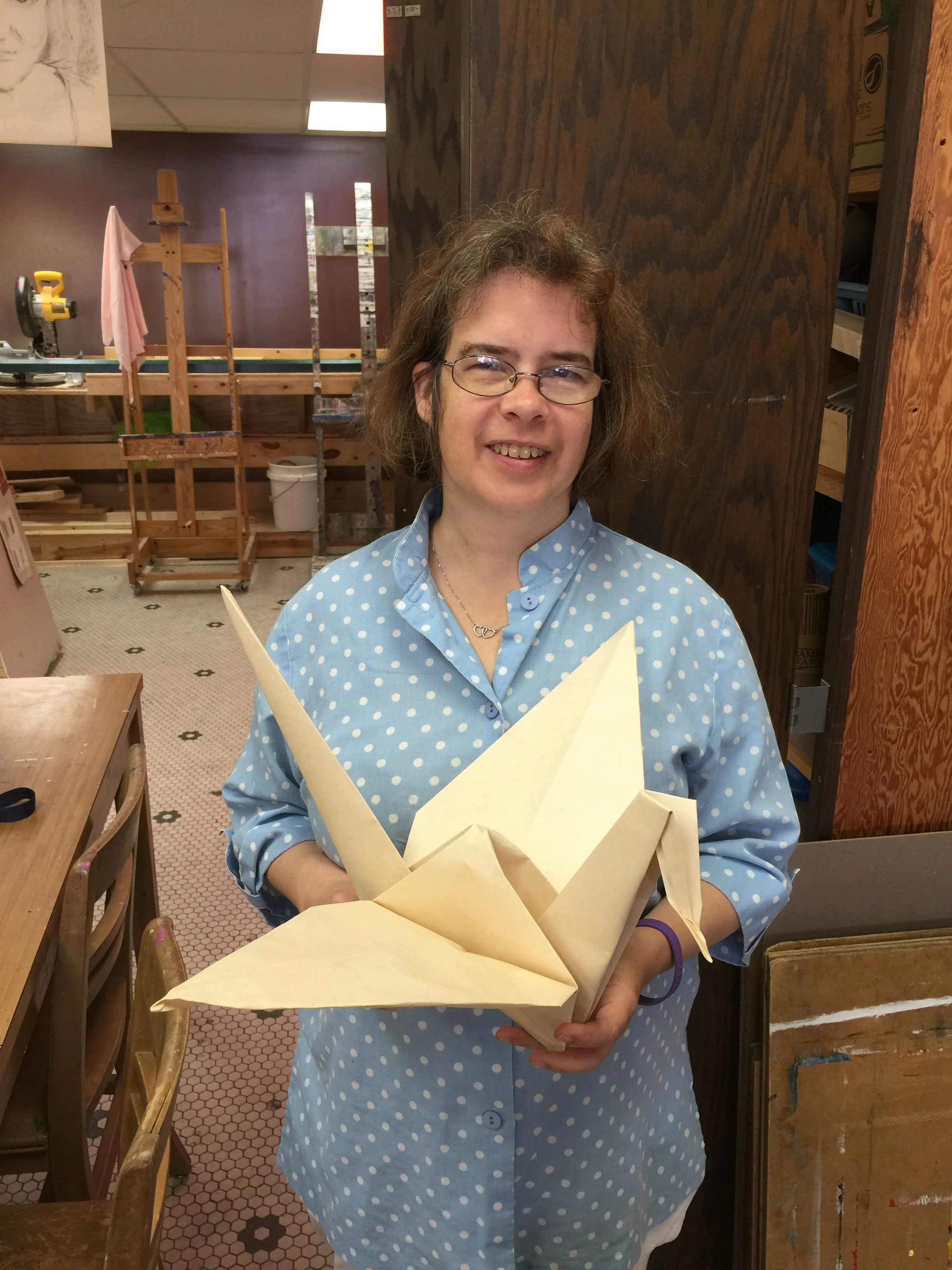 A woman with glasses and curly hair holding a folded paper sculpture in an art studio or workshop.