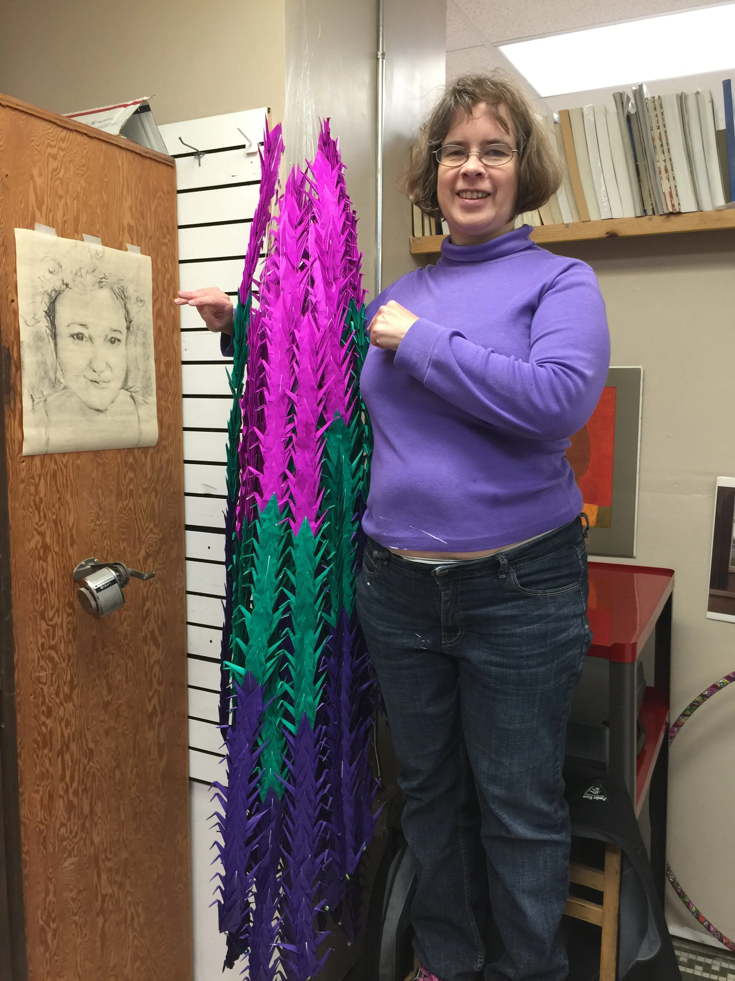 A woman with glasses and curly hair smiling, standing indoors next to colorful origami paper sculptures resembling tall, spiky plants.